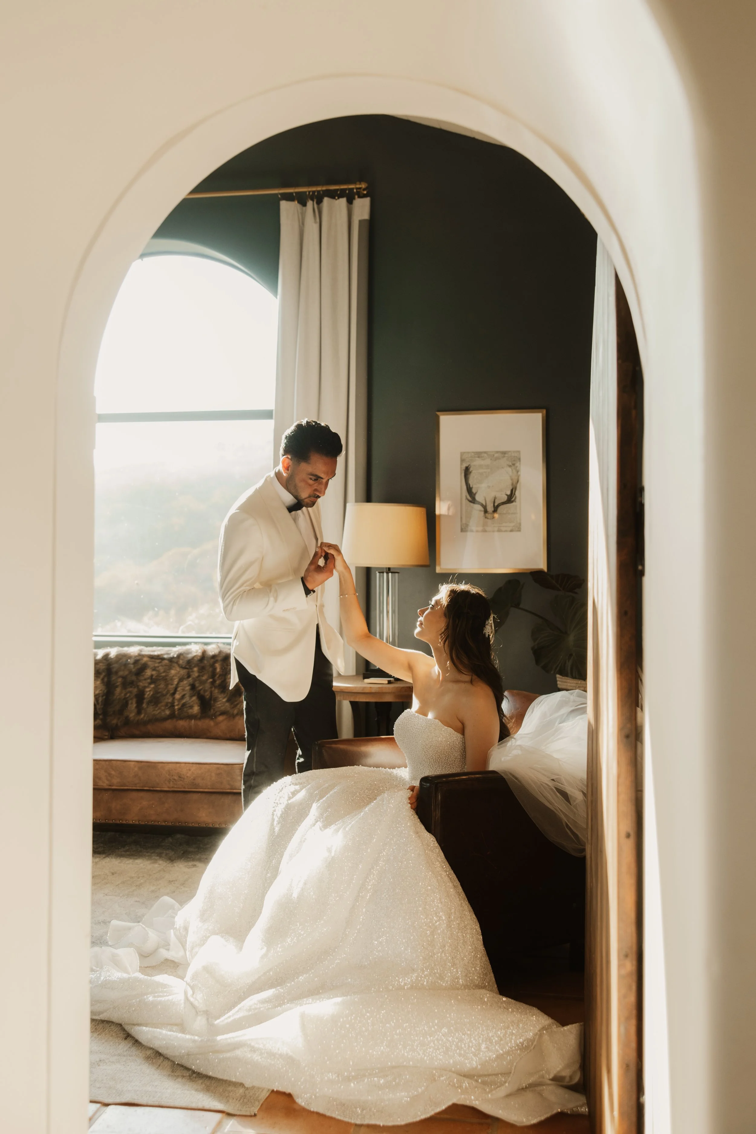 A bride in a white wedding gown sitting on a cushioned chair in a hotel room, with a groom standing in front of her, holding her hand, in front of a large window with sunlight streaming in, a dark-colored wall with framed pictures, and a couch with a furry blanket.