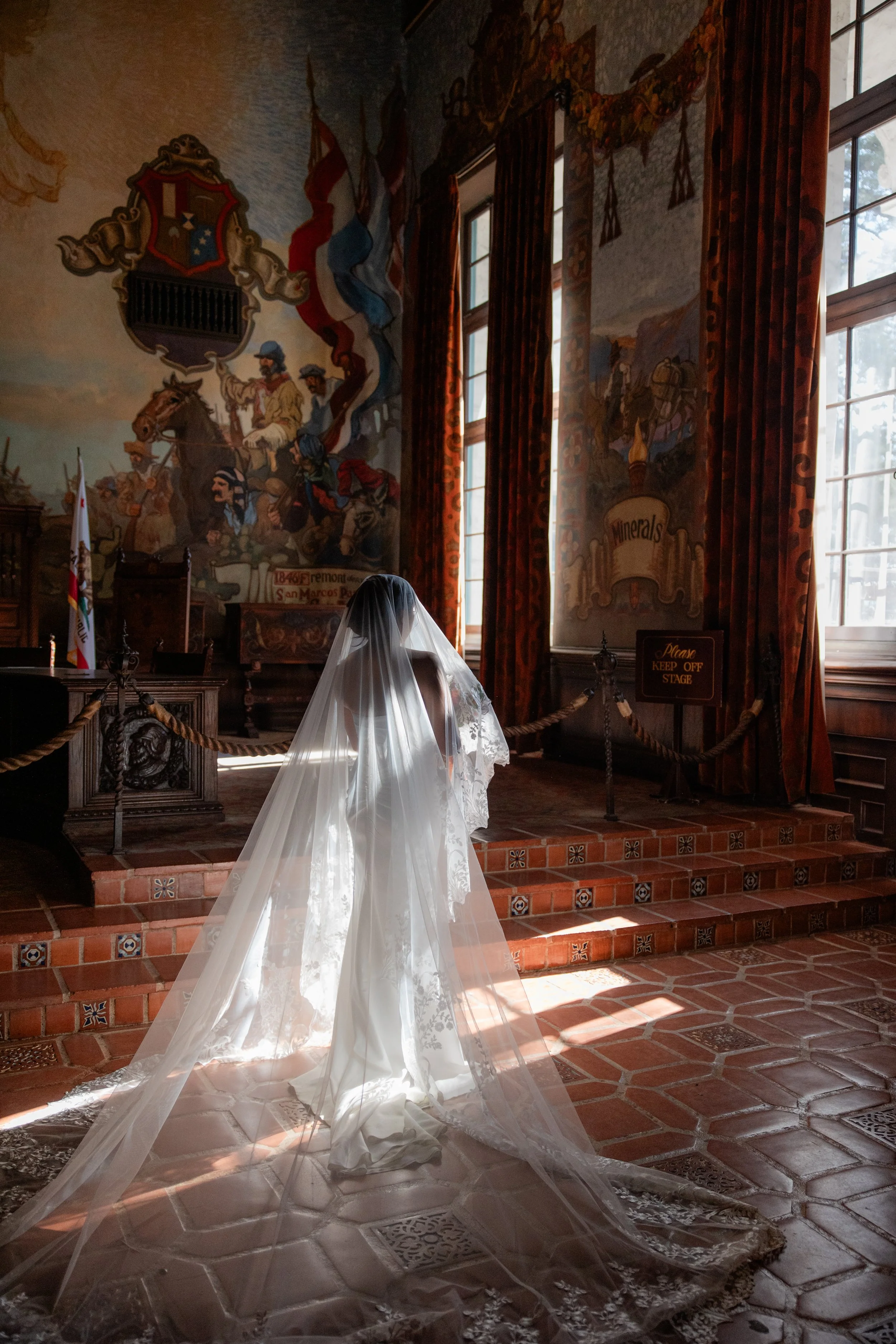 A woman dressed in a white wedding gown, with a long train and veil, standing inside a historic church with wooden floors, large windows, and decorated walls and drapes.