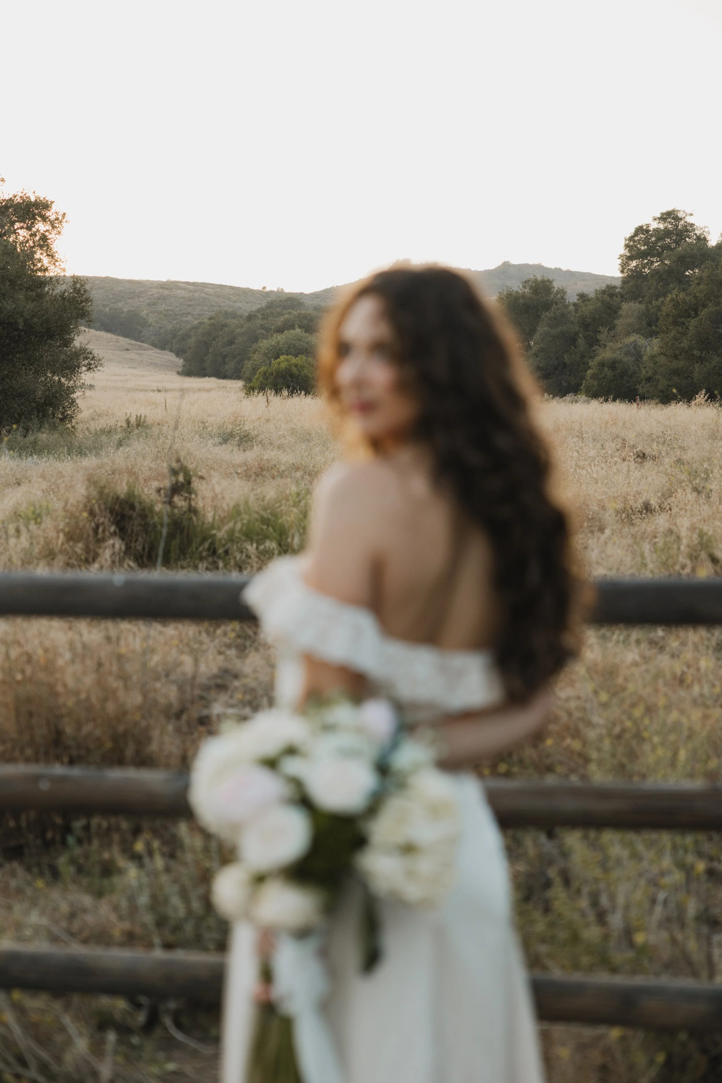 Blurred bride in a white dress sitting on a park bench with a bouquet of white flowers, in an open field with trees and hills in the background during sunset for her golden hour irvine engagement photoshooot