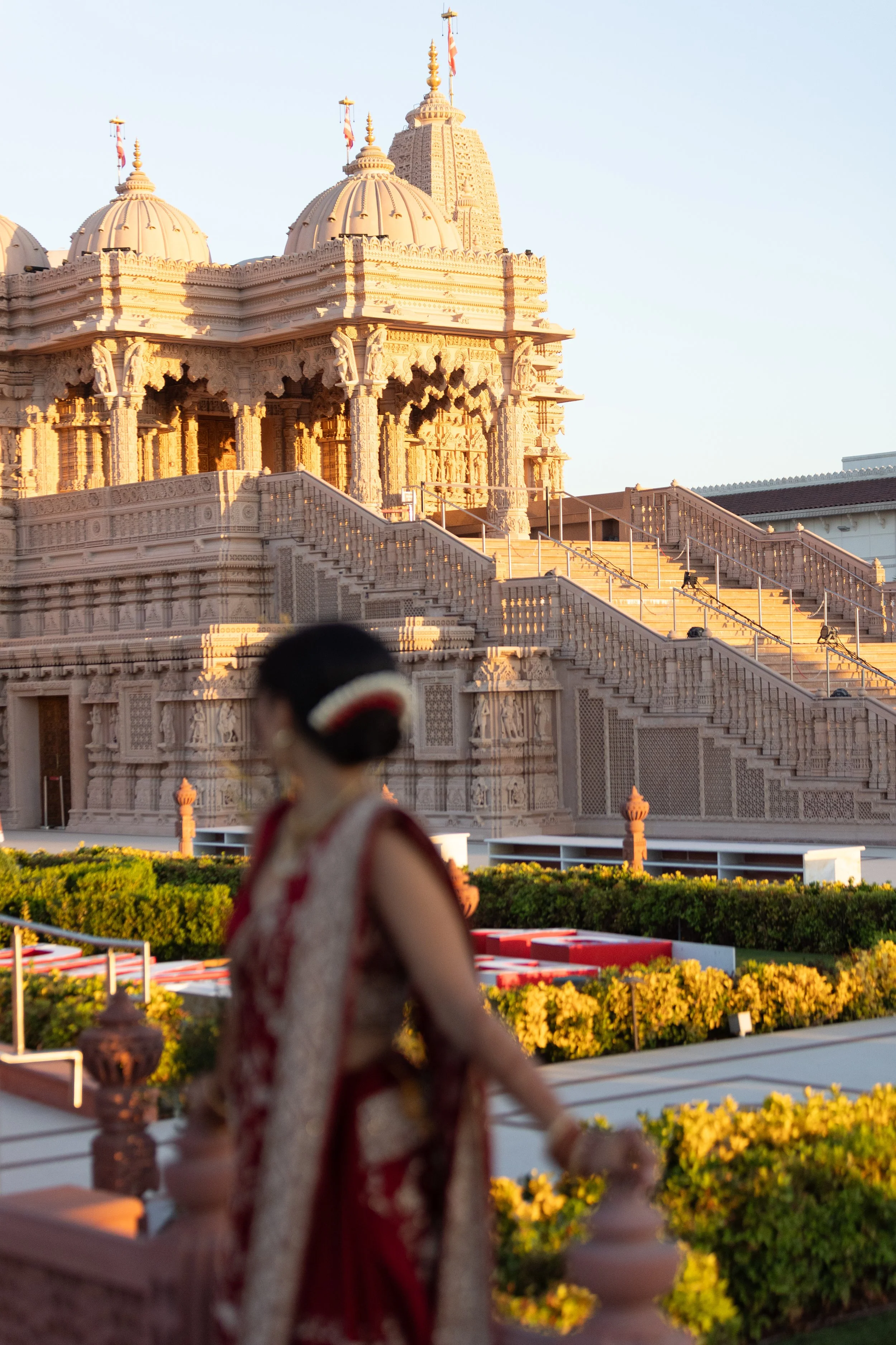 A temple with intricate carvings and multiple domes, set against a clear sky, with steps leading up to its entrance. Blurred figure of a woman in traditional attire is in the foreground.
