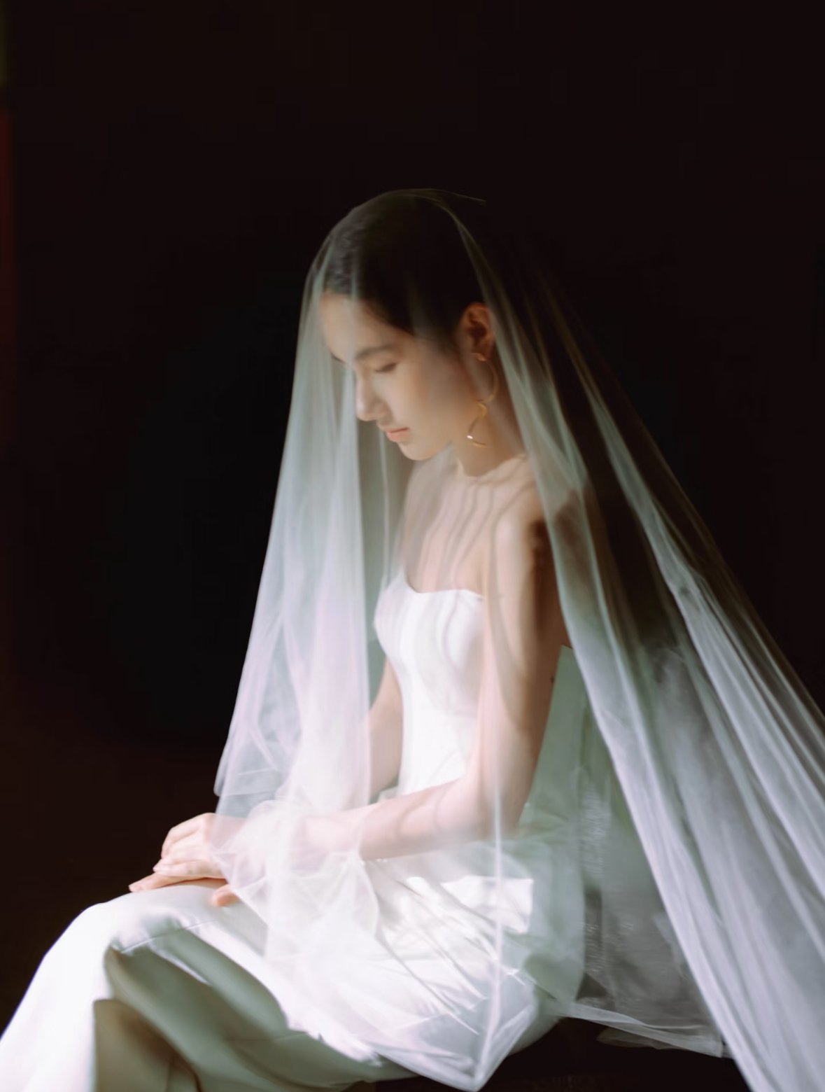 A woman dressed in a white wedding gown with a veil, sitting against a black background with her head bowed and eyes closed.