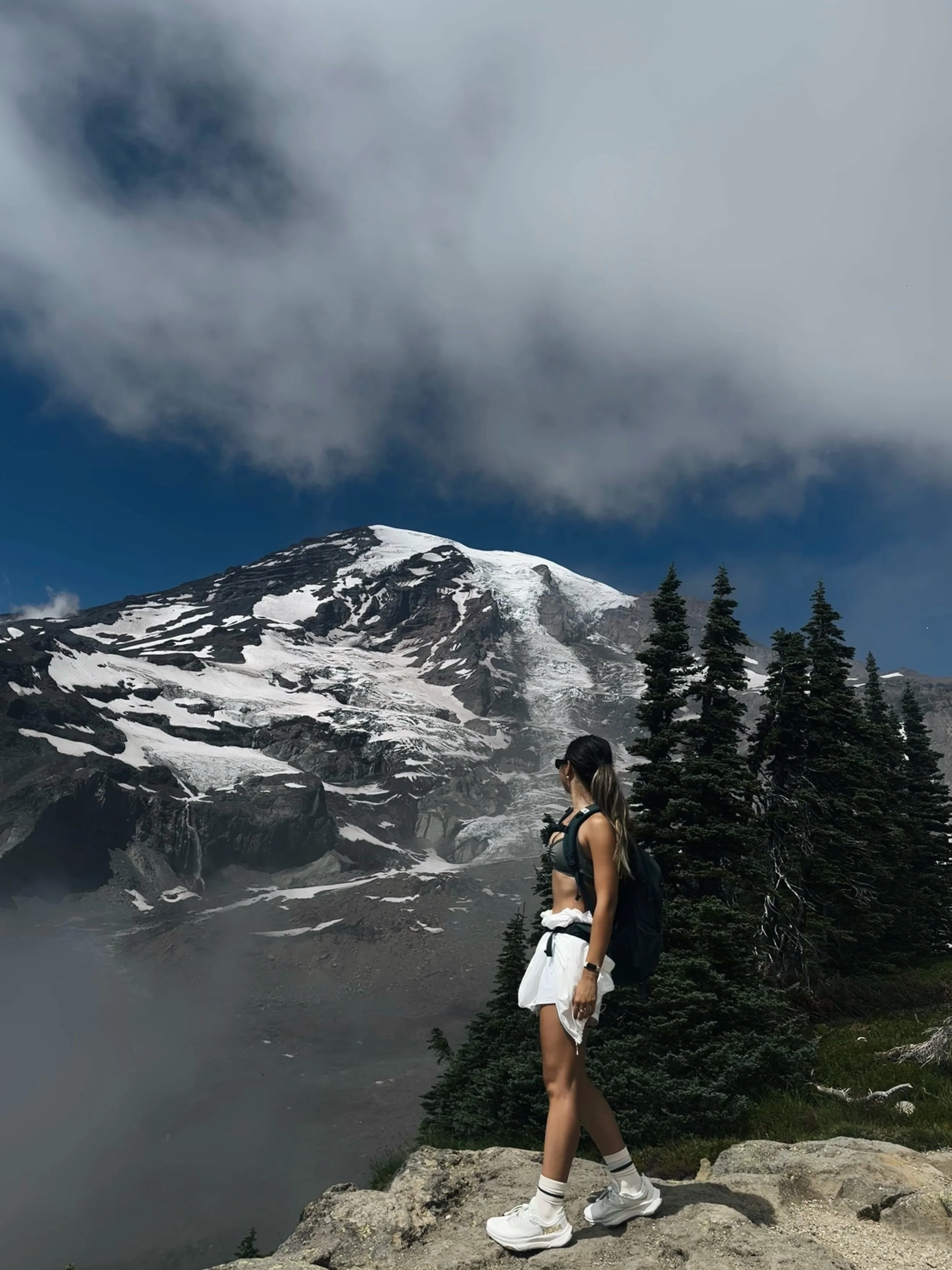 A woman stands on a rocky ledge overlooking a mountain with snow and ice, surrounded by trees under a cloudy sky.