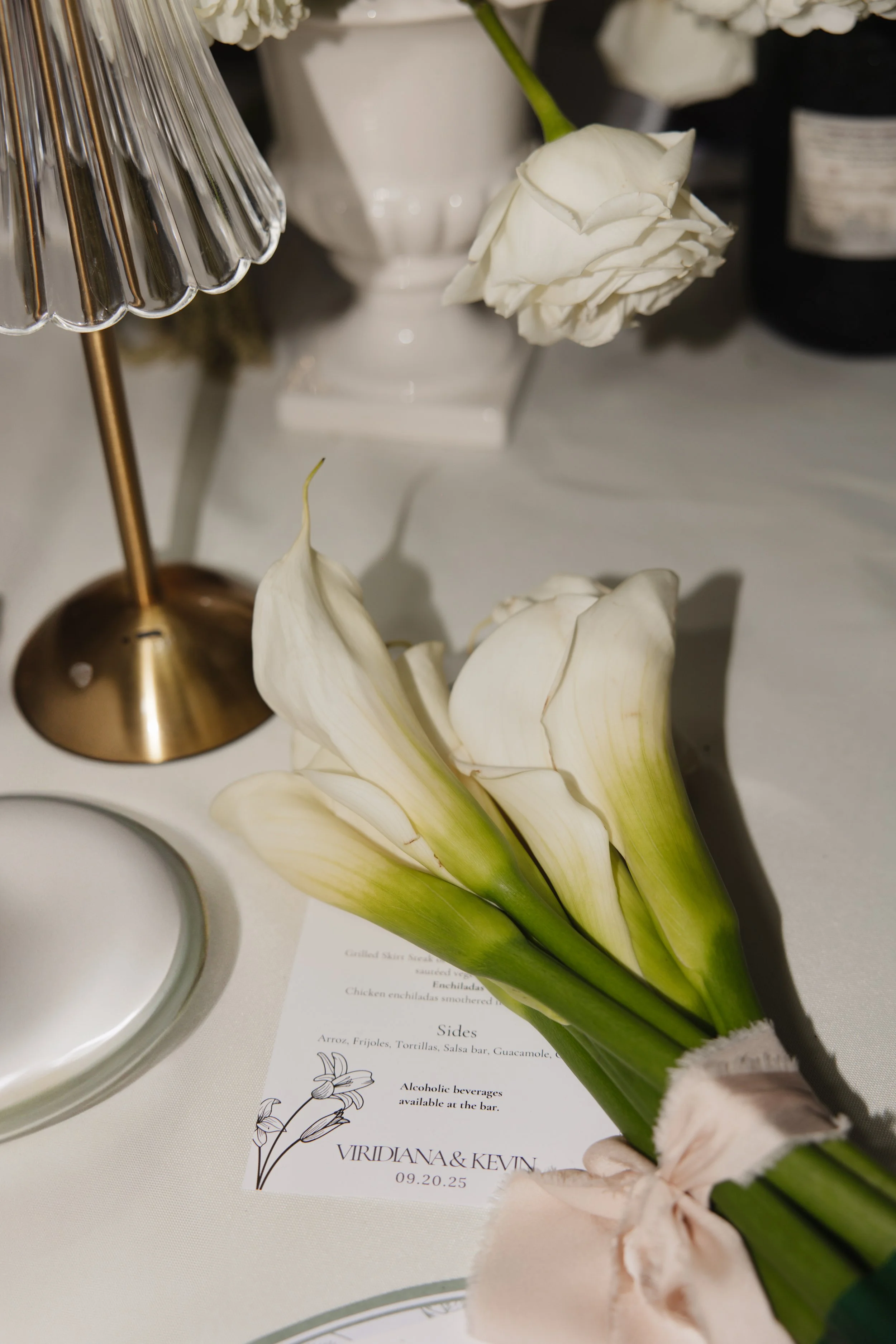A white vase with white flowers and green stems, a gold-colored lamp, and a white candle holder on a white surface with a printed menu and small decorative items.
