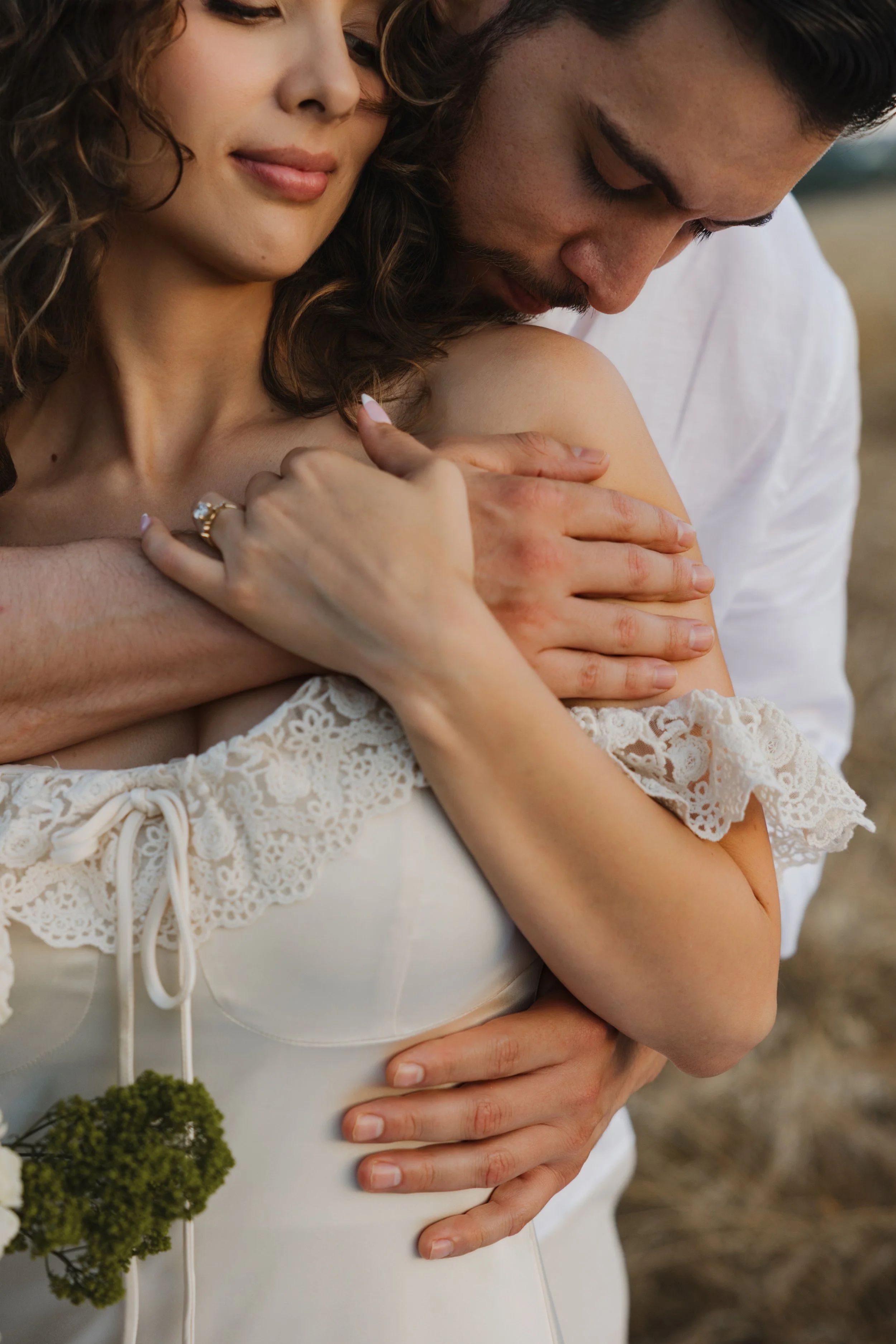 A couple hugging outdoors, with a woman in a white lace dress and a man in a white shirt, embracing closely with their arms around each other.
