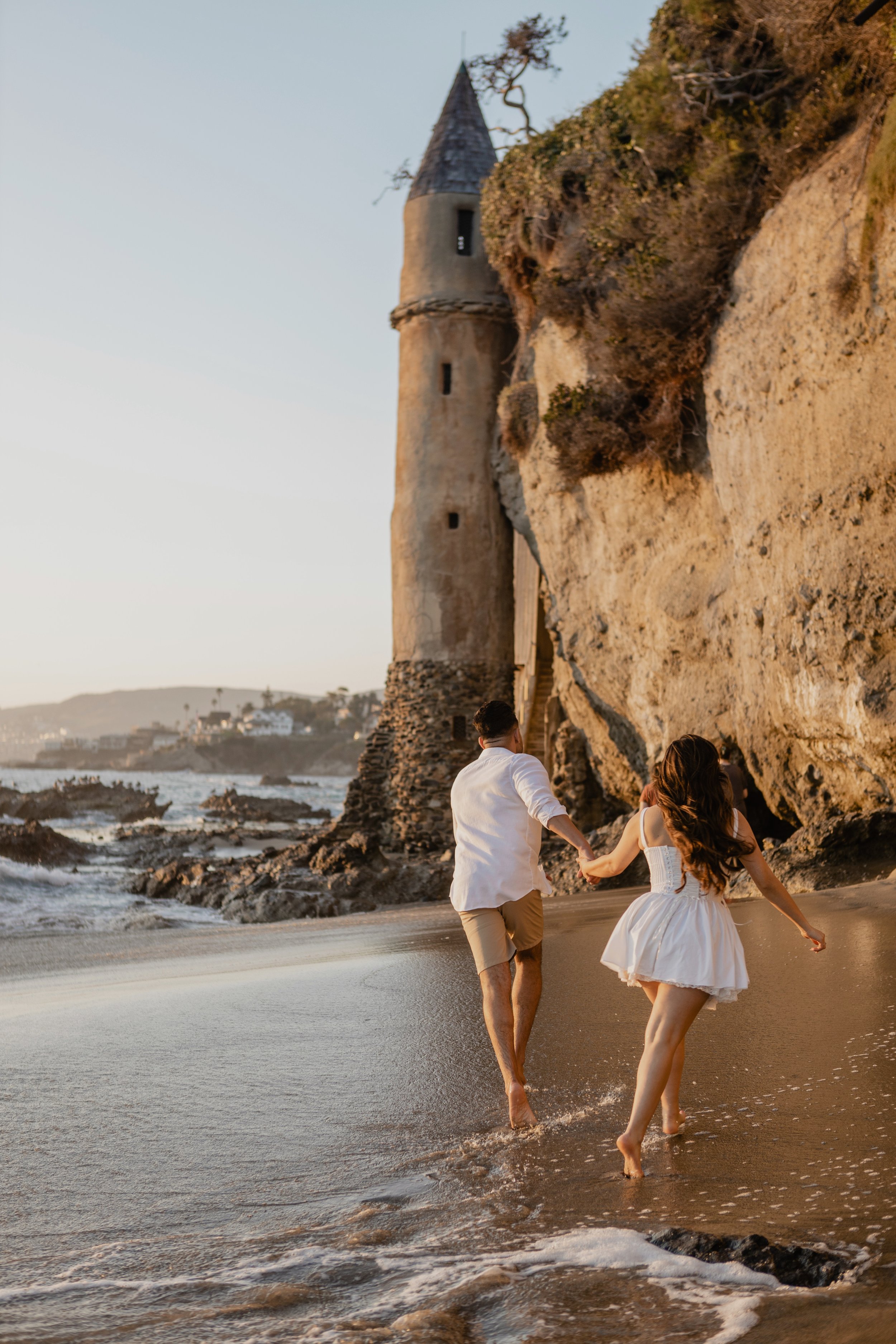 A couple holding hands and running along the beach at sunset, with a historic tower on a rocky bluff in the background for their laguna beach engagement photos