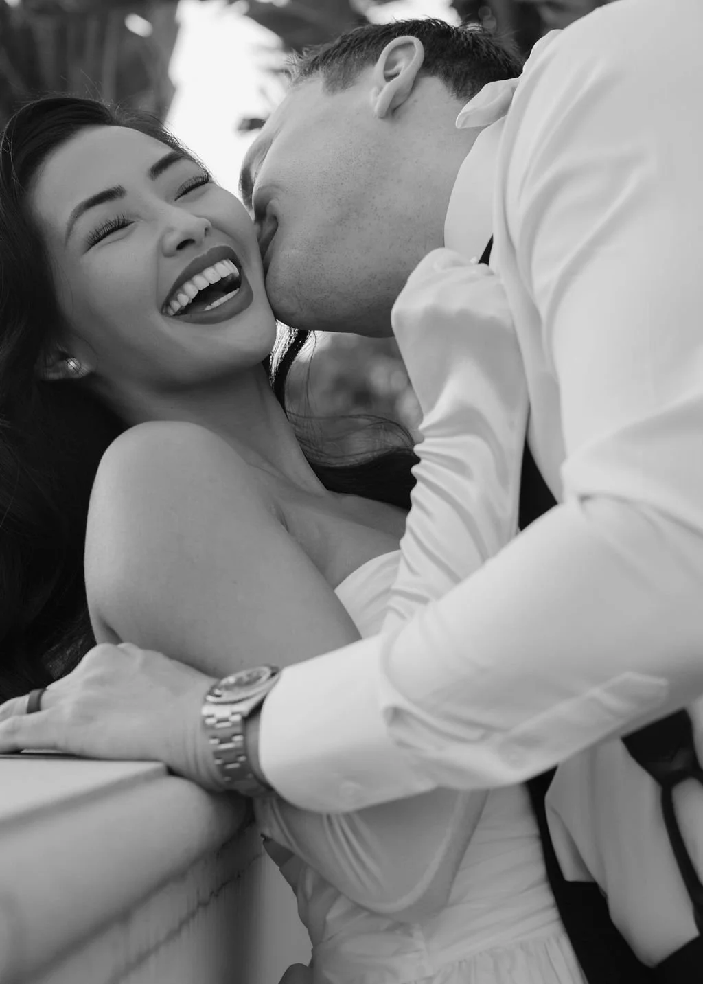 A woman with dark hair smiling and a man kissing her cheek in a black and white photo.