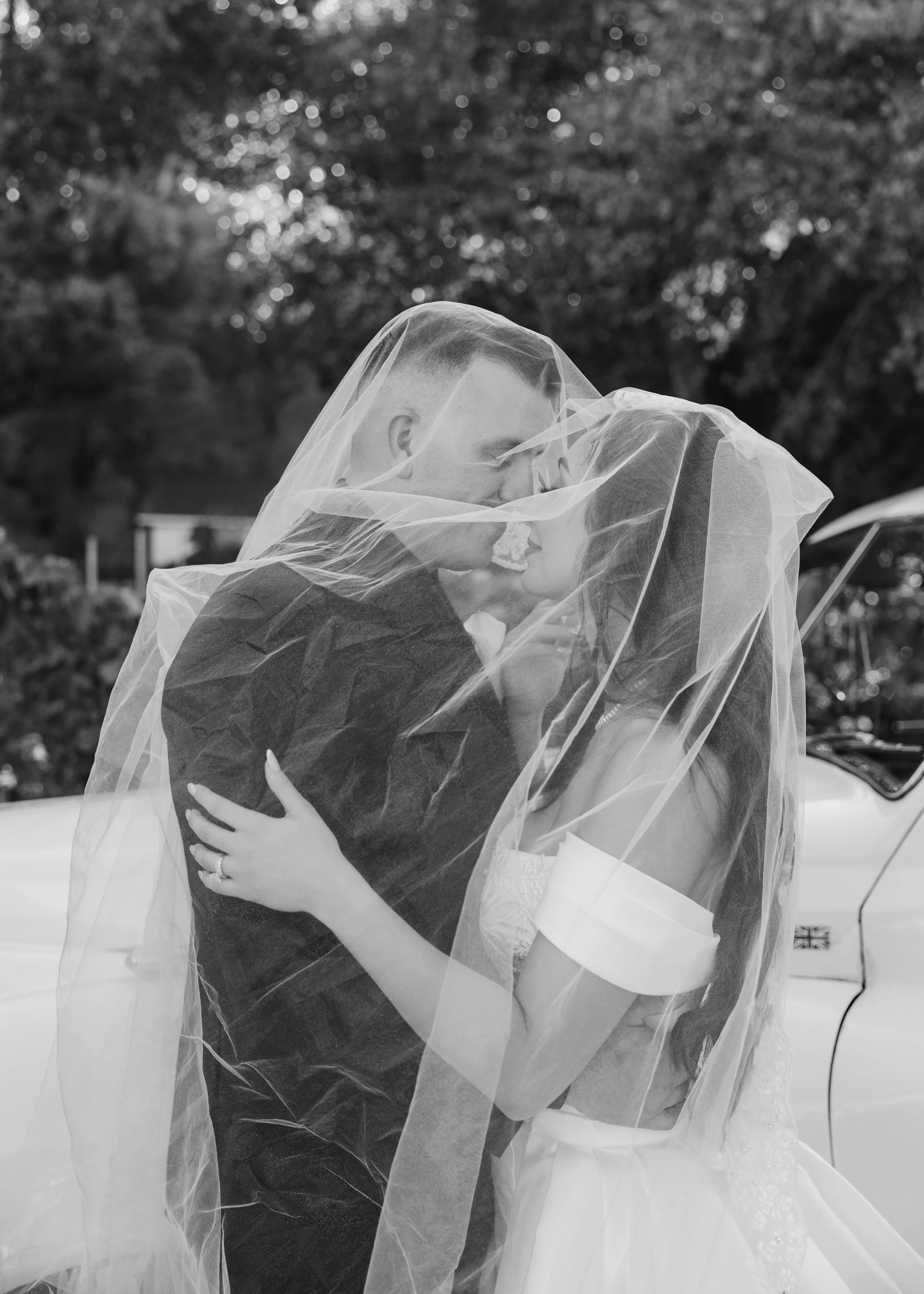 A bride and groom embracing and kissing, with the bride wearing a veil and wedding dress, and the groom in a suit and tie, outside near a car with trees in the background.