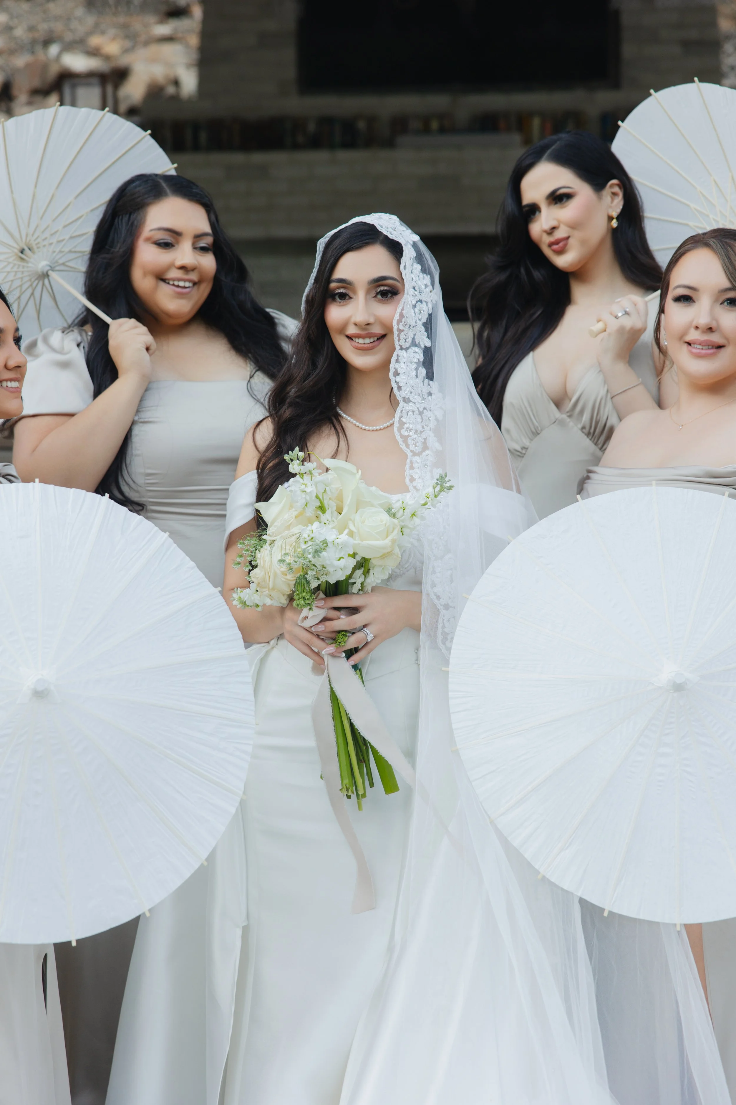 A bride holding a bouquet of white flowers and standing amid her bridesmaids, who are holding white parasols during a wedding celebration.