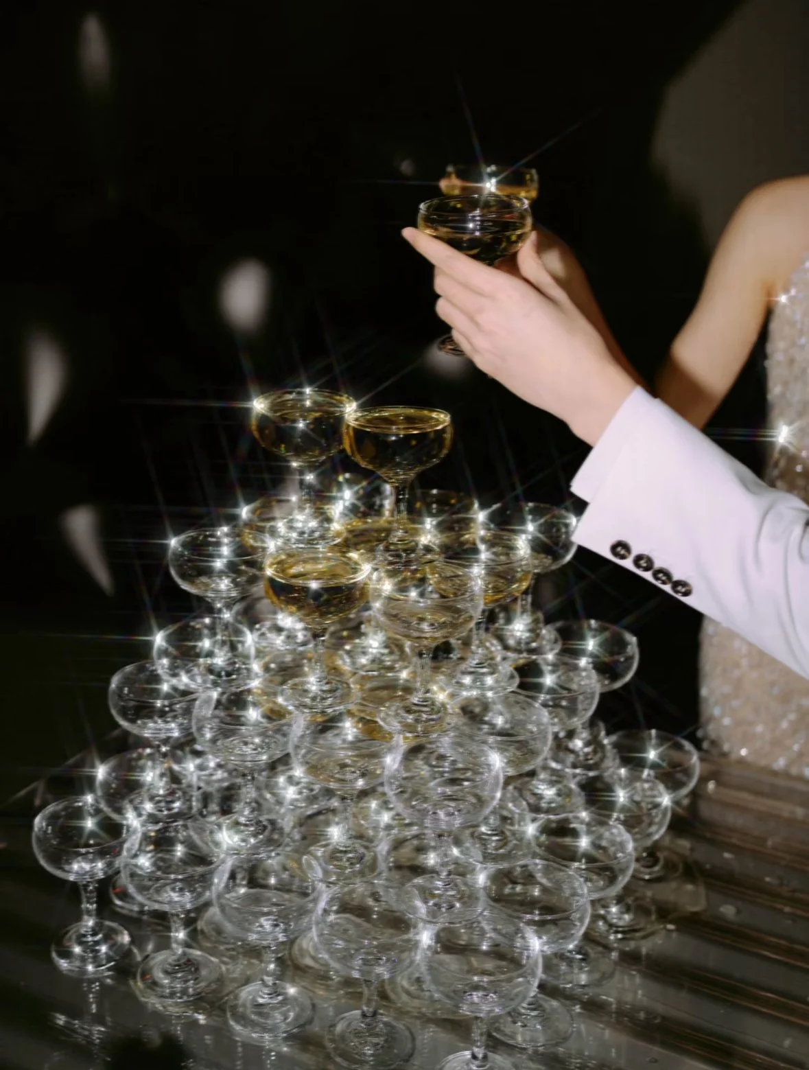 A pyramid of champagne glasses with some glasses filled with champagne and a person in a white suit placing a glass at the top. The scene is decorated with sparkling light effects.