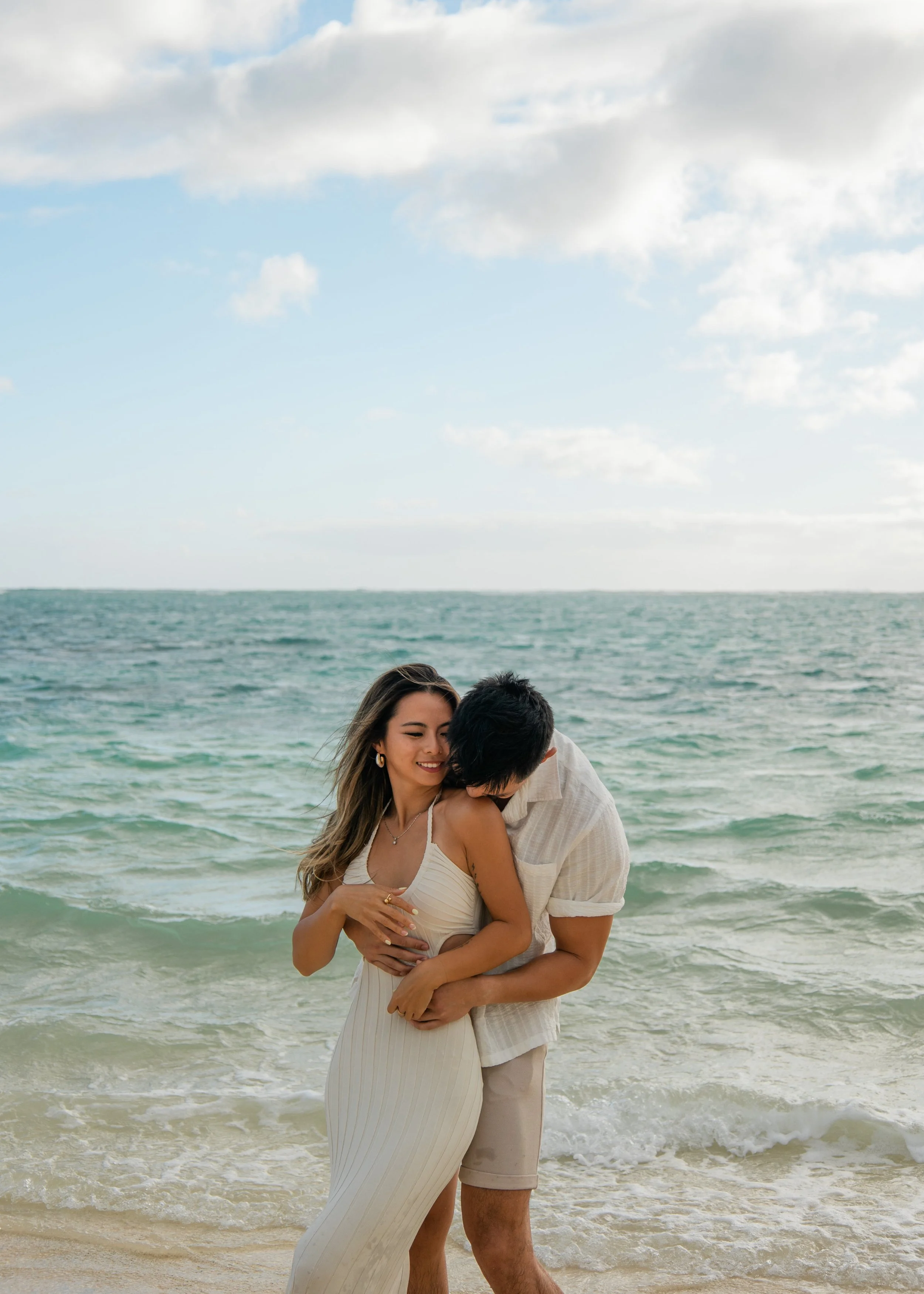 A couple embracing on a beach with ocean waves and cloudy sky in the background.