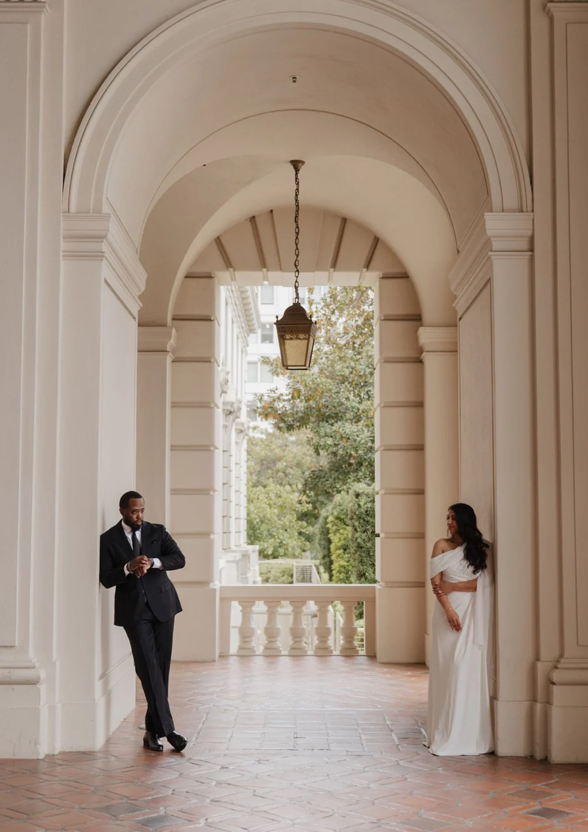 A man in a black suit and a woman in an elegant white gown stand opposite each other in a grand, covered outdoor corridor with archways and a hanging lantern. The corridor overlooks a garden with trees.