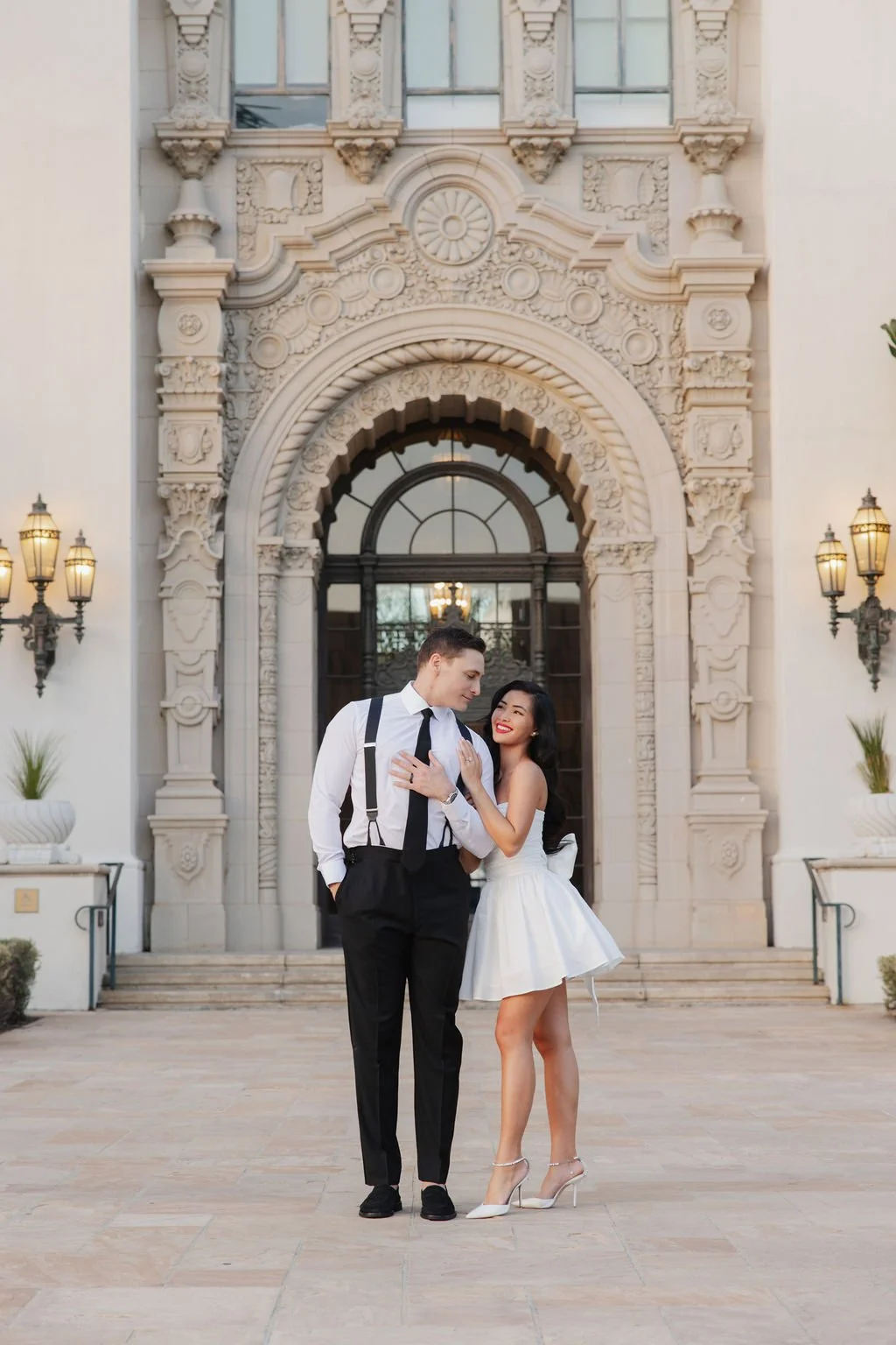 Bride and groom dressed elegantly standing in front of a historic ornate building entrance, smiling and looking at each other, with the woman touching the man's chest, both appearing joyful for their civil ceremony photoshoot.