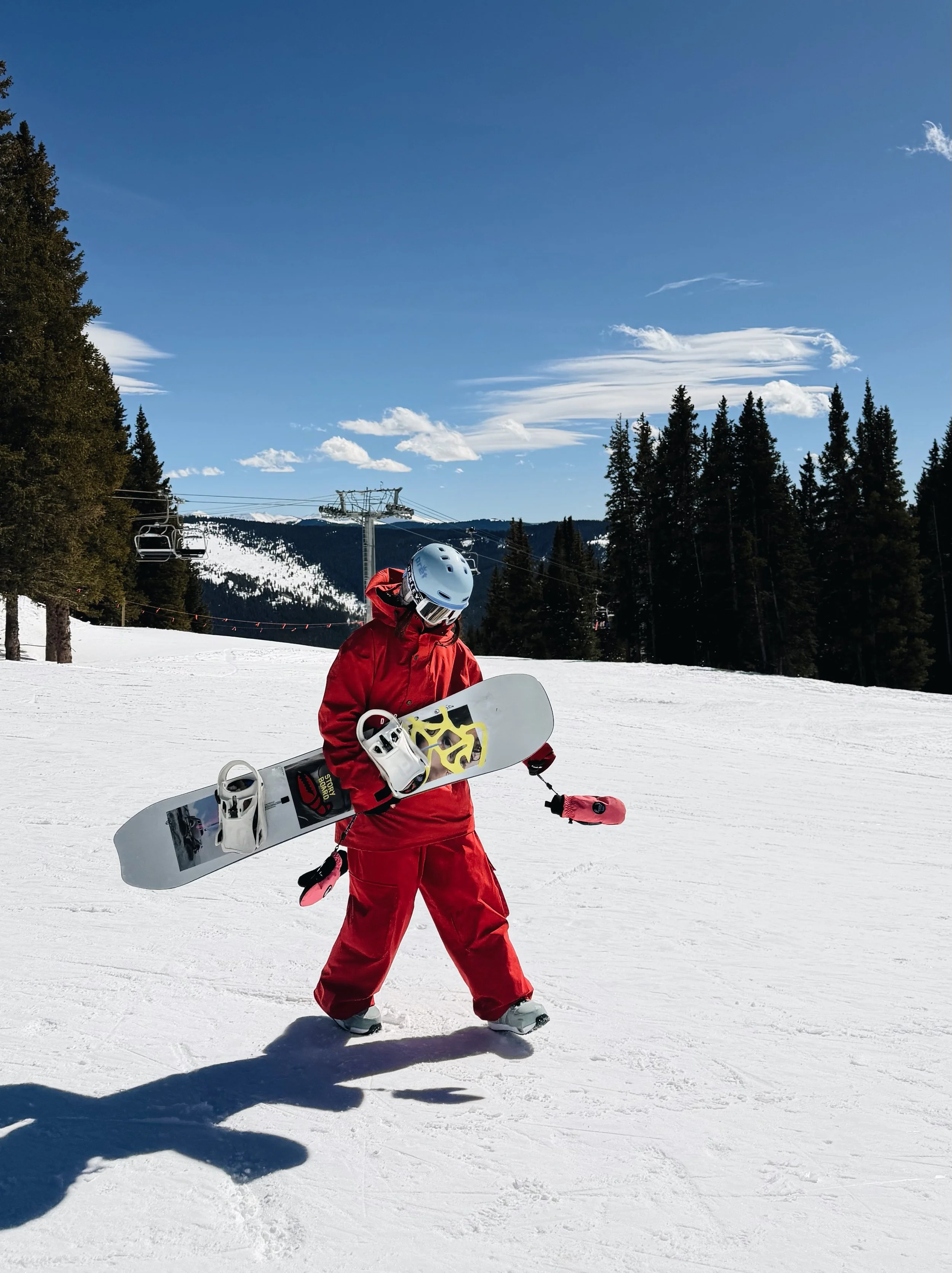 A person dressed in red snowboarding gear is walking on a snowy slope while holding a snowboard and wearing a helmet and goggles. Pine trees and ski lifts are visible in the background.