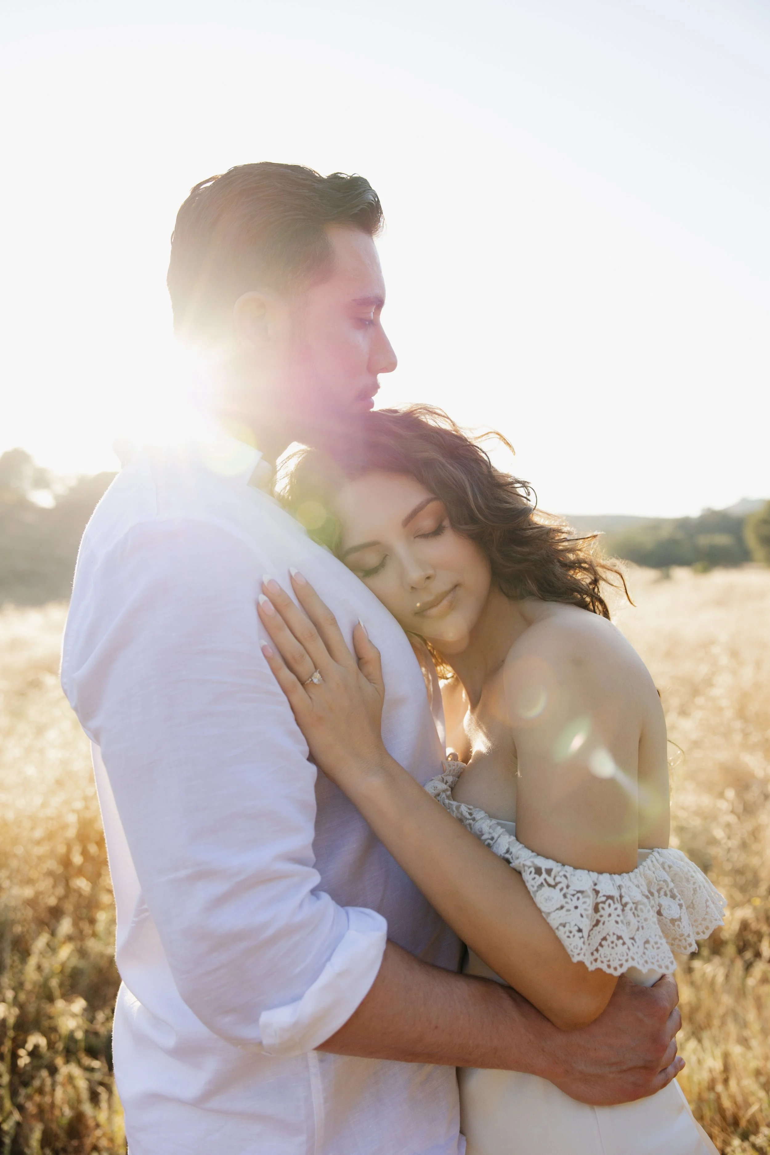 A couple embracing in a field during sunset, with the woman resting her head on the man's chest and both with serene expressions.