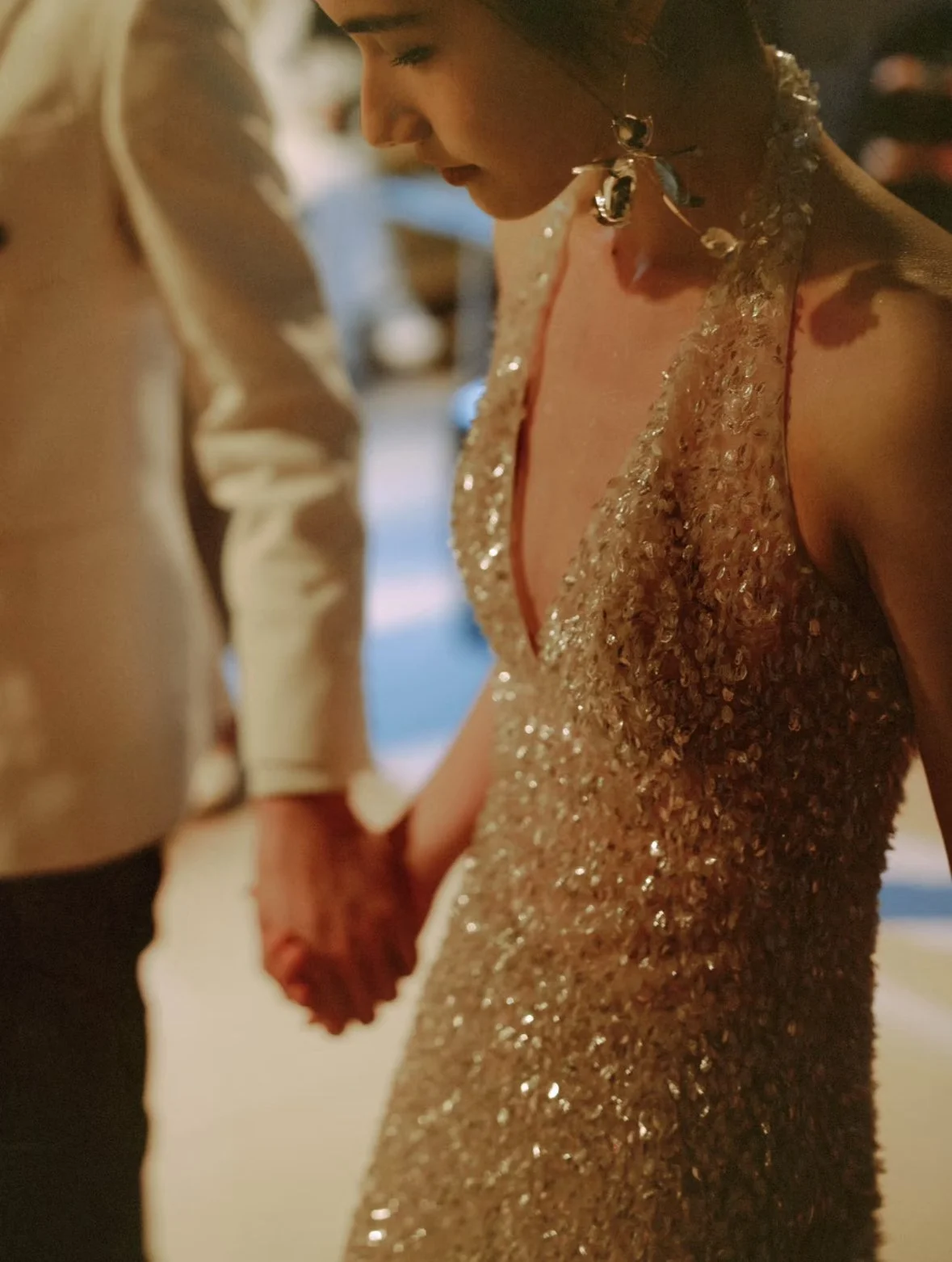 A close-up of a woman wearing a sparkly, sequined dress and jewelry, holding hands with a man, in a dimly lit setting, possibly a wedding or formal event.