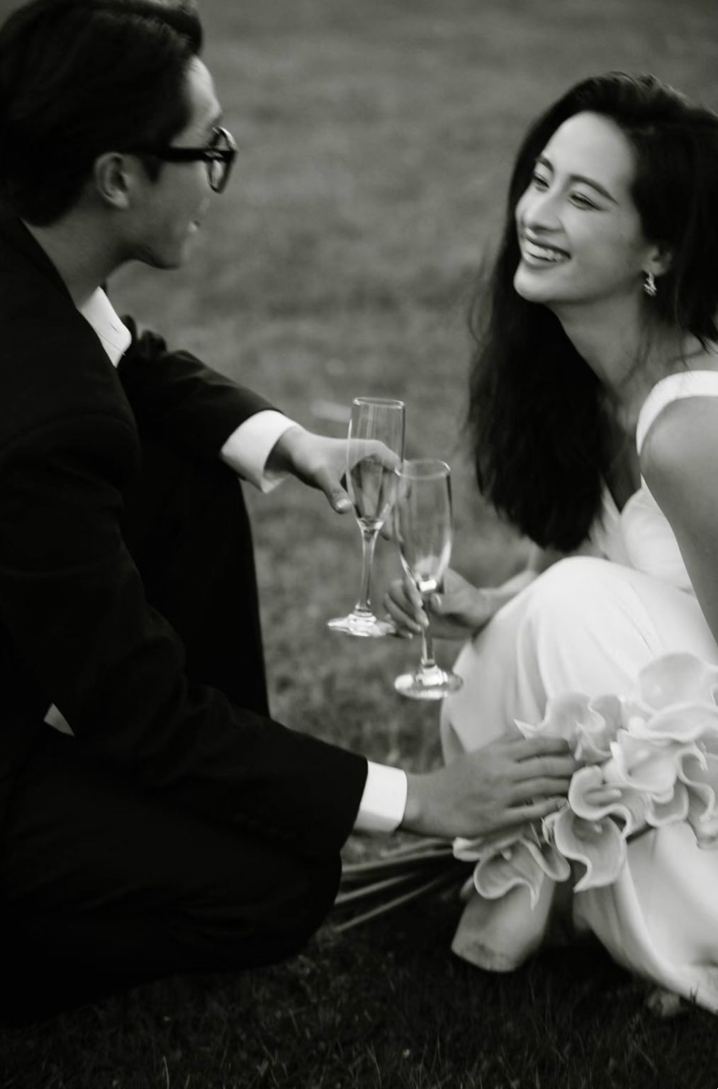 A black and white photo of a man and woman smiling and toasting with champagne glasses at an outdoor event, with the woman seated on the grass.
