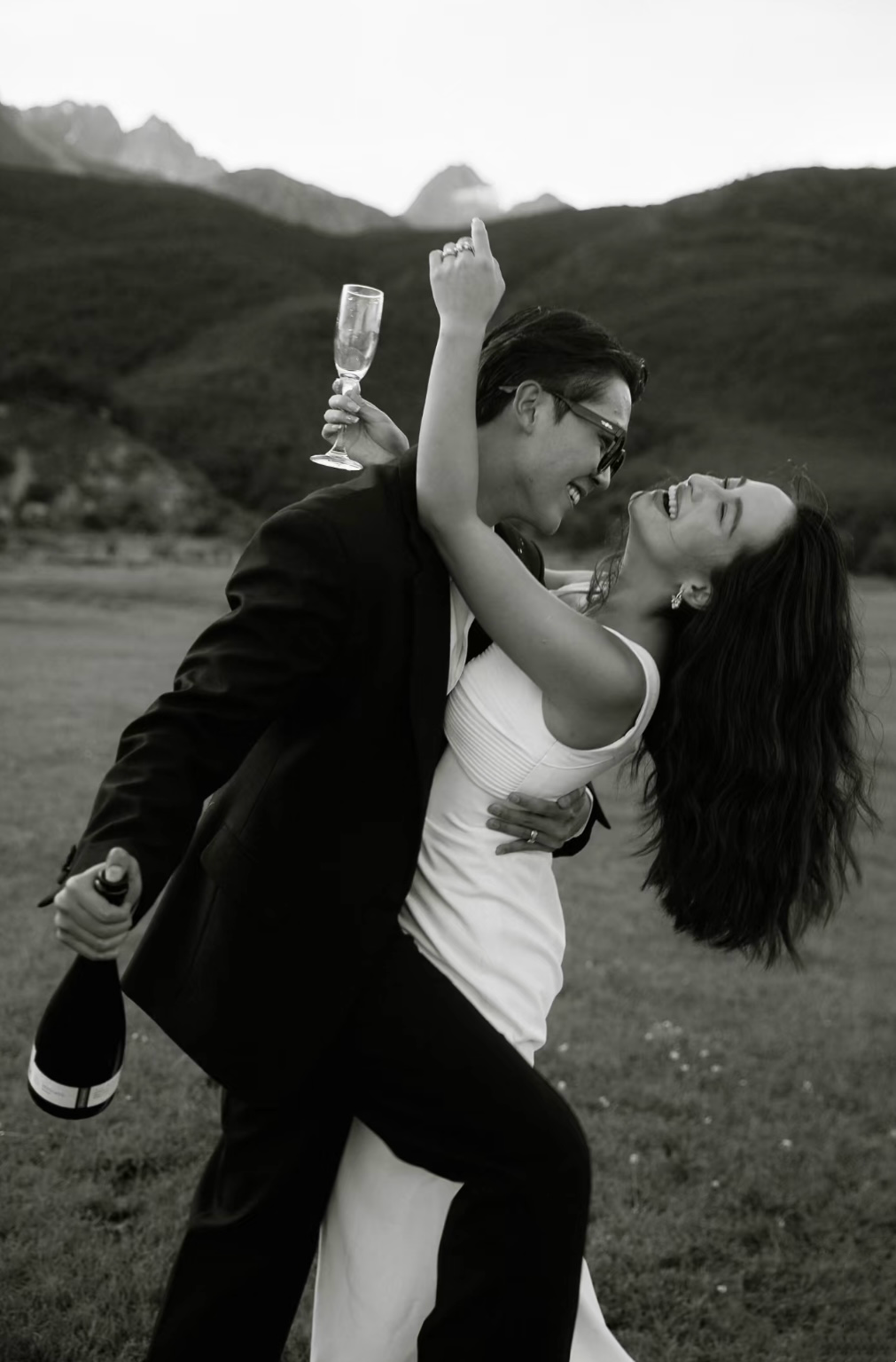 A couple dressed in formal attire celebrating outdoors, with the man in a tuxedo and the woman in a white dress, holding a champagne bottle and glass, smiling and laughing in a mountain landscape.