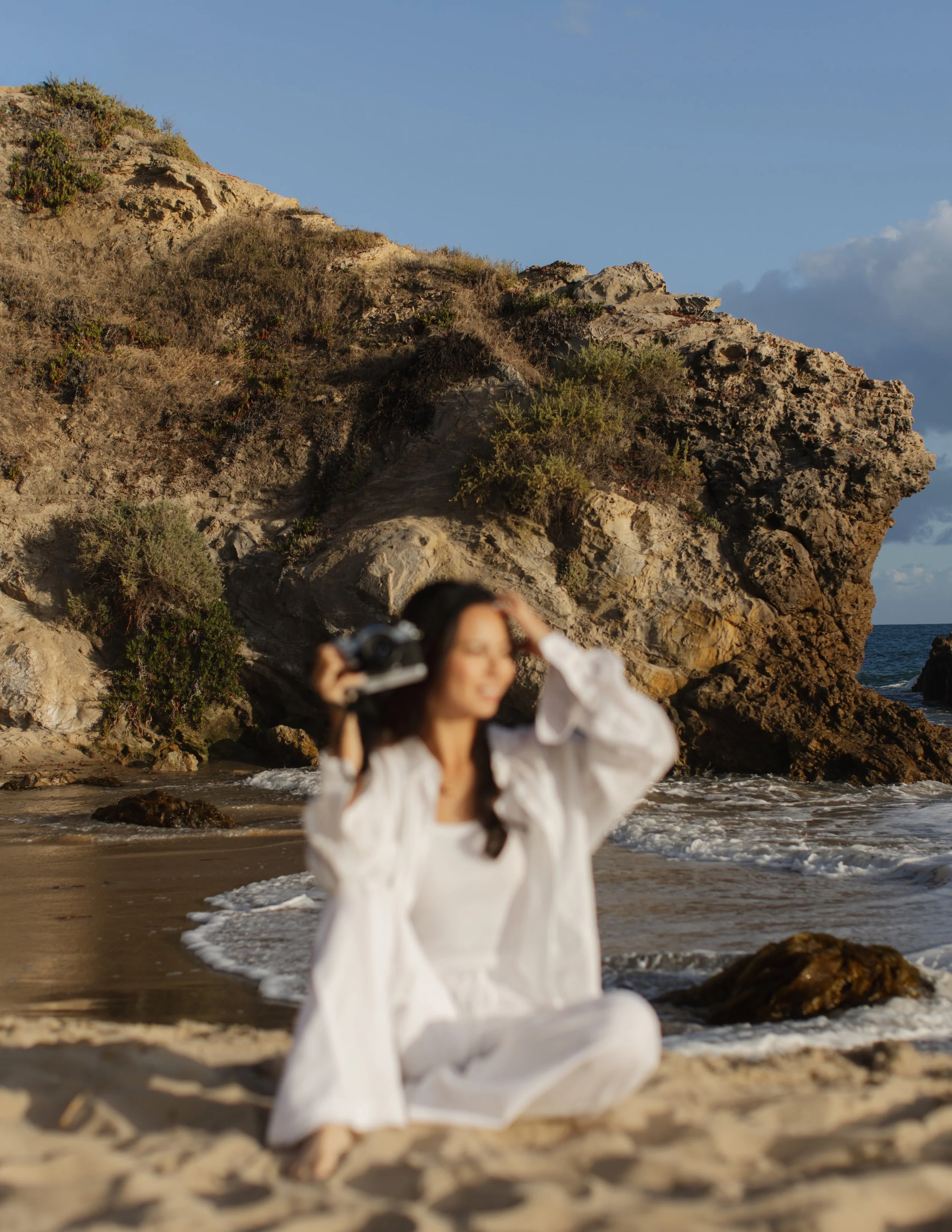 A woman in an all-white outfit sitting on a sandy beach, holding a camera to her eye, with rocky cliffs and blue sky in the background.