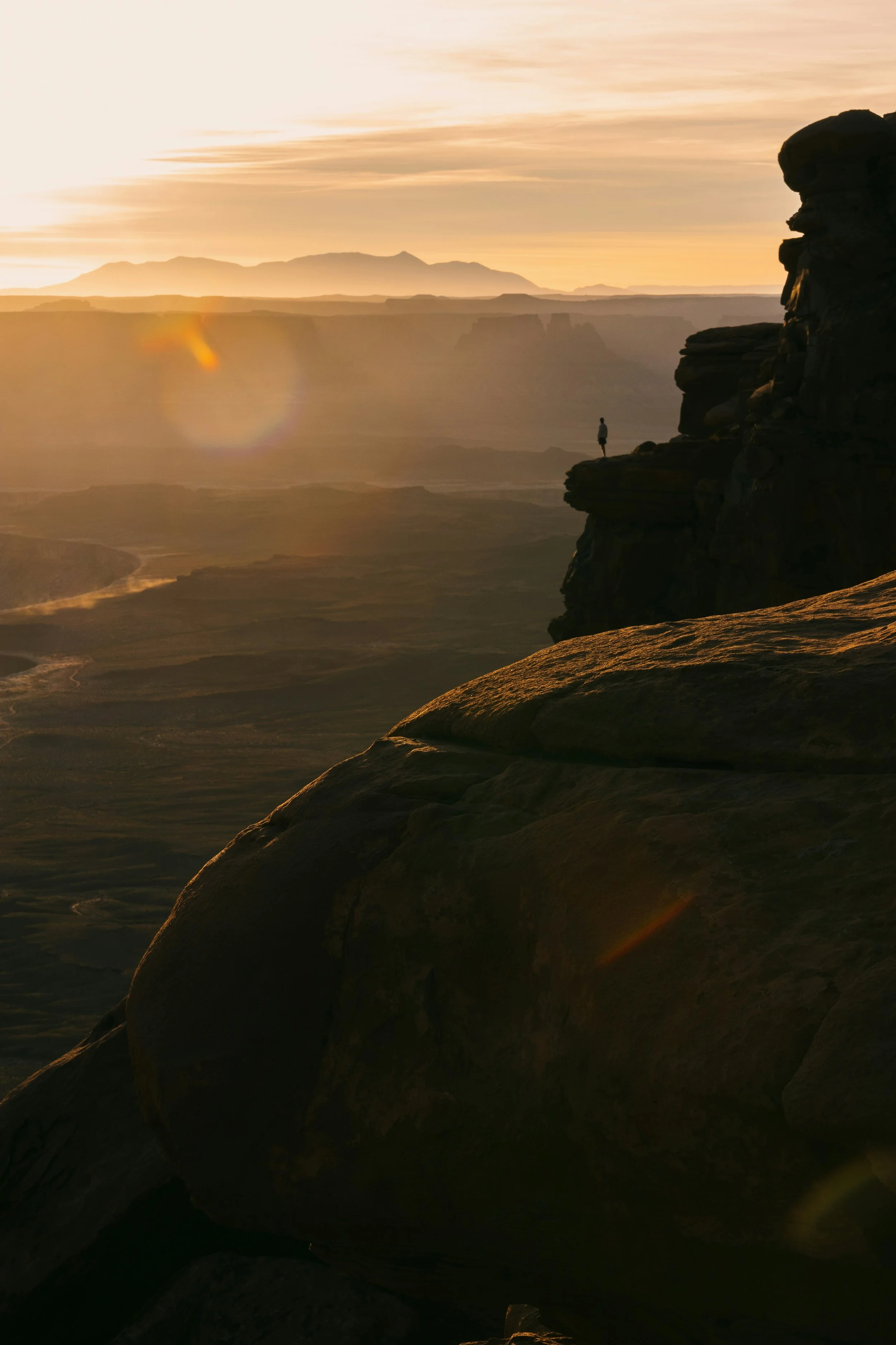 A person standing on a rocky ledge overlooking a canyon and mountains during a golden sunset.