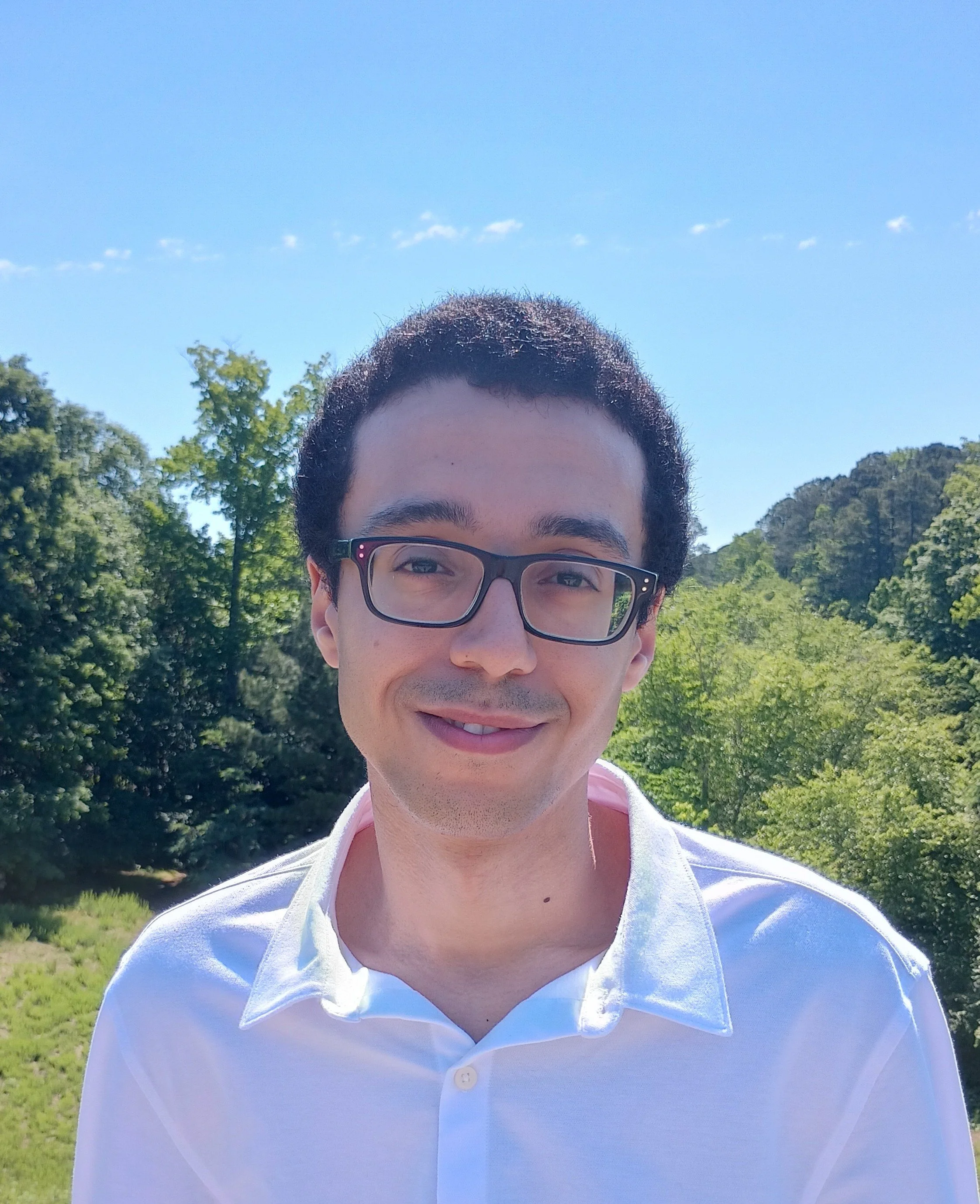 Close-up of a young man with dark hair and glasses, smiling outdoors with trees and blue sky in the background.