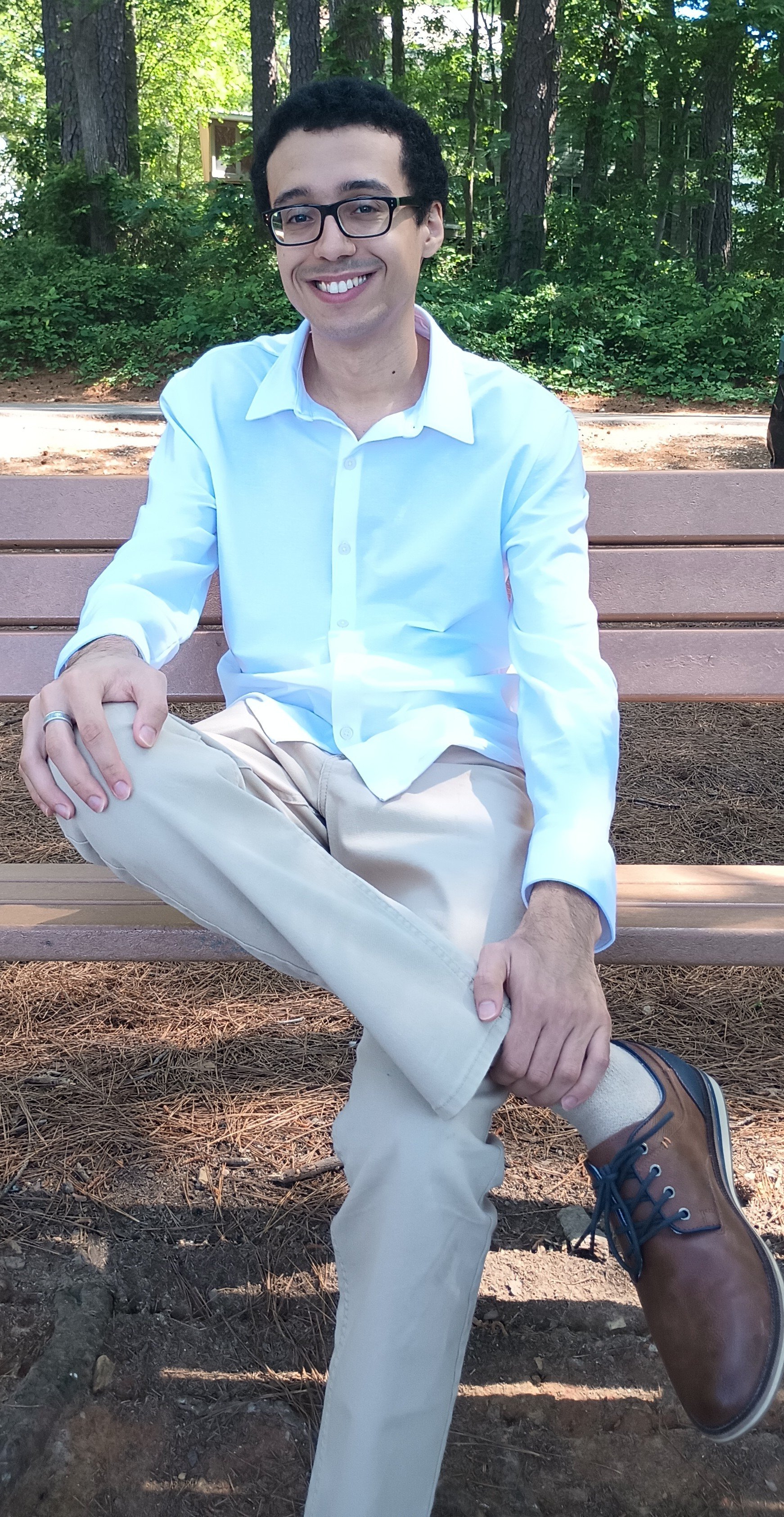 A young man with glasses and dark curly hair is sitting on a wooden park bench, smiling, outdoors with green trees in the background, wearing a white long-sleeve shirt, beige pants, and brown shoes.