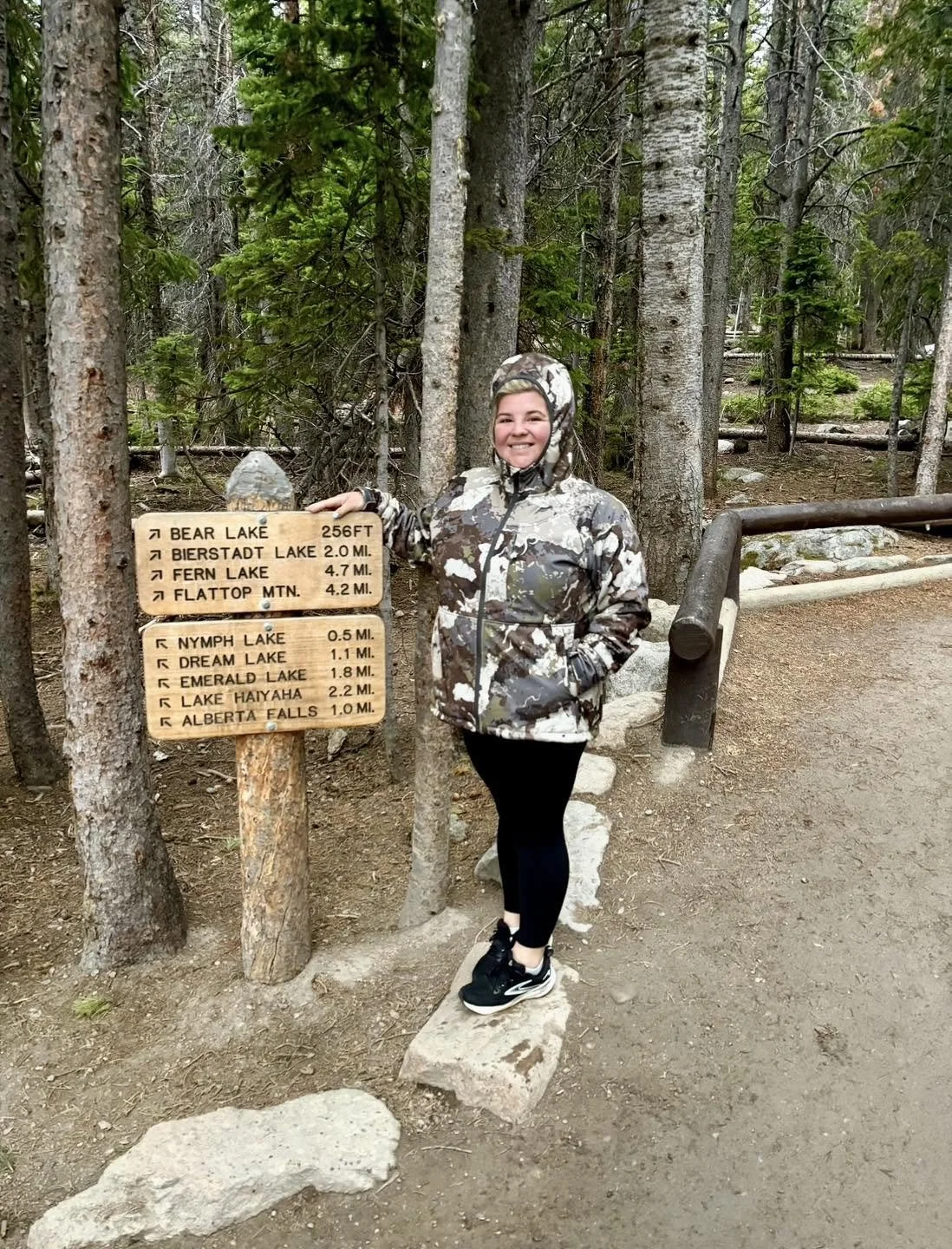 A smiling woman in camouflage jacket and black leggings standing next to a wooden trail sign in a forested area. The sign indicates directions and distances to various lakes and geographic features.