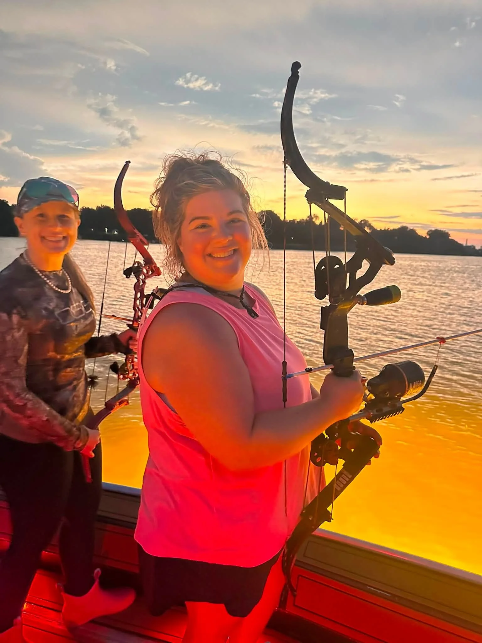 Two women stand on a boat during sunset, fishing with a bow and arrow fishing set, one woman holding a bow and smiling, water and trees in the background.