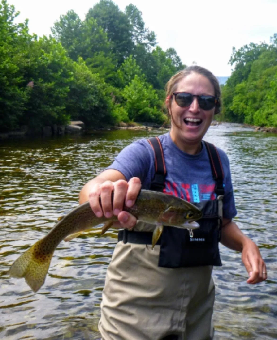 Woman in sunglasses smiling holding a fish in a river with green trees in the background.