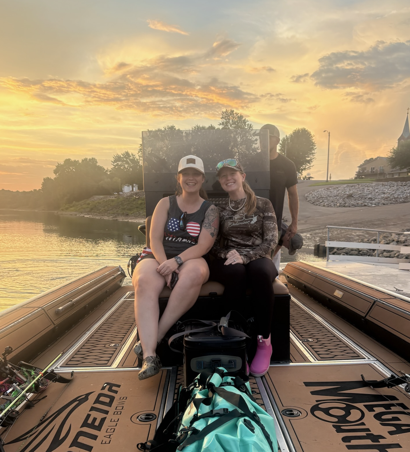 Two women sitting on a boat docked on a river during sunset, smiling at the camera, with another person standing behind them. The scene includes a beautiful sky with orange and yellow hues, trees, rocks, and houses along the riverbank.