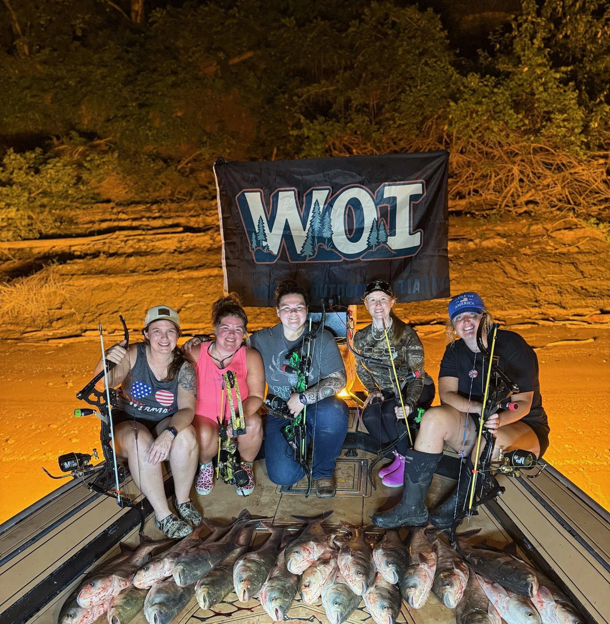 Group of people posing with fishing gear in front of a pile of fish on the floor, with a wooded area and a flag with trees in the background at night.