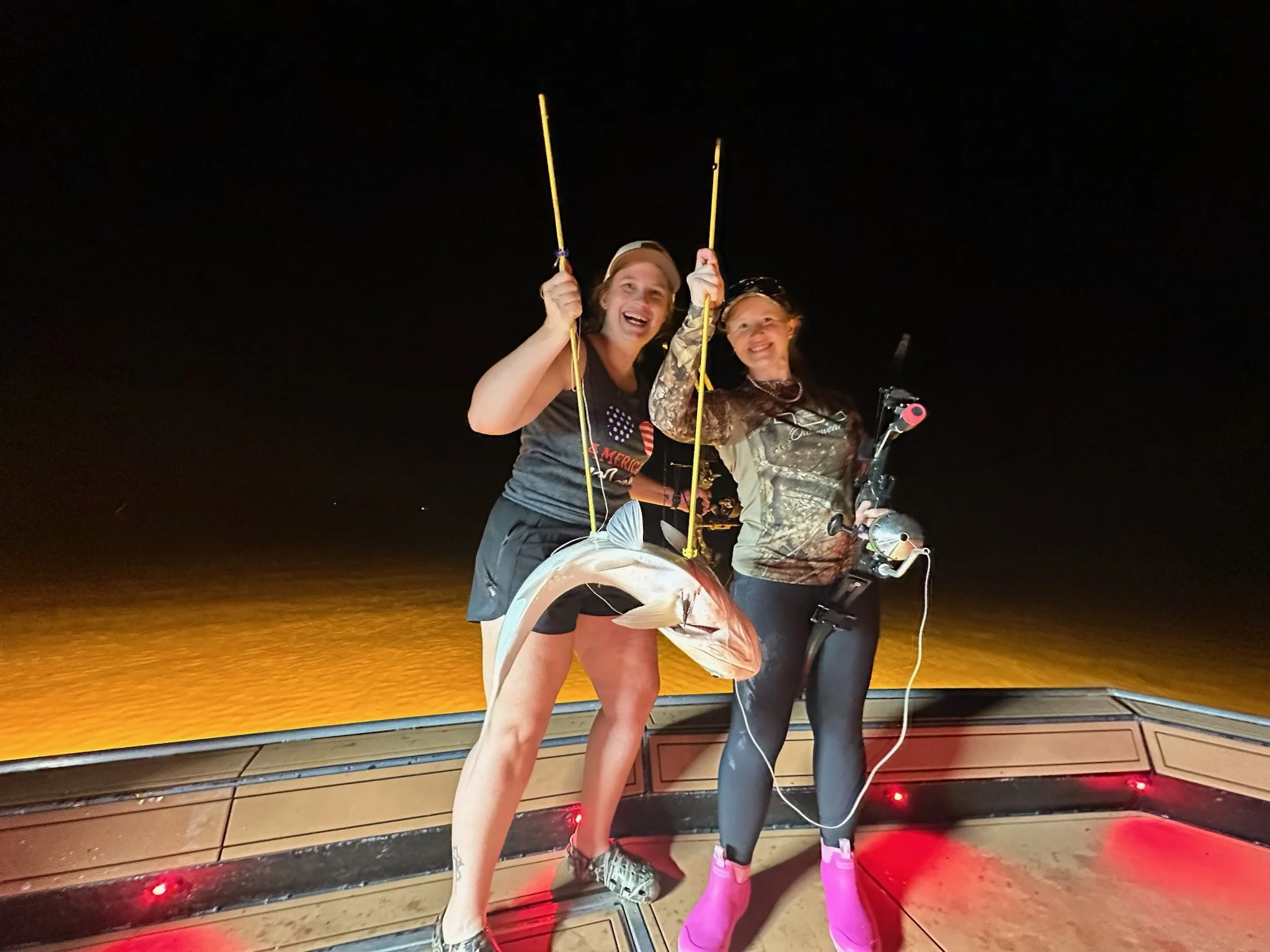 Two women smiling and holding fishing rods on a boat at night with a fish on the deck, illuminated by red and yellow lights.