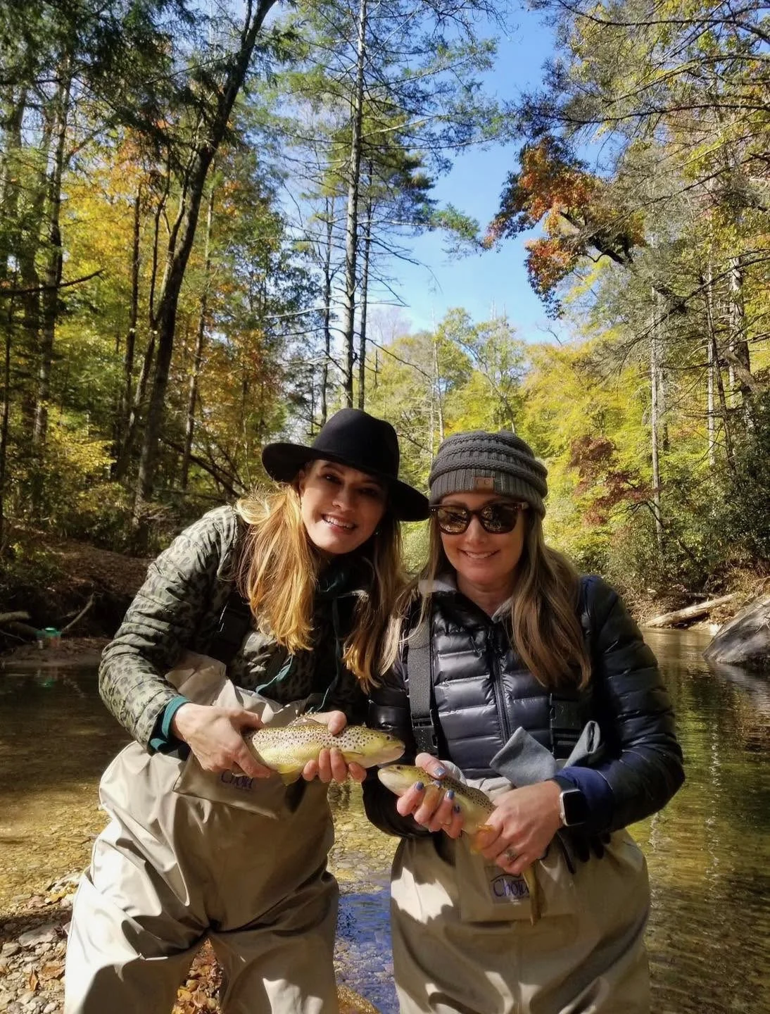 Two women in fishing gear holding fish in a creek surrounded by trees with fall foliage.