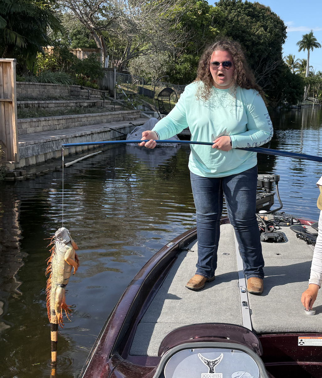 A woman with curly hair wearing sunglasses and a light blue long-sleeve shirt holding a fishing rod on a boat, with a fish caught on the line over a canal with trees and stairs in the background.