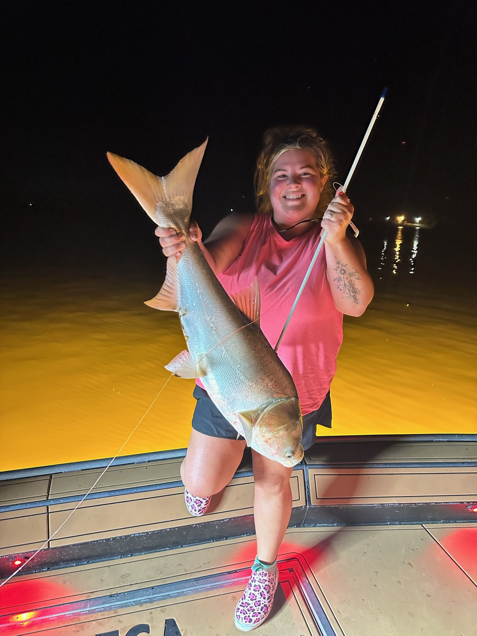 A woman smiling, holding a large fish with a yellow tail at night. She is wearing a pink sleeveless shirt, shorts, and animal print rain boots. She is standing on a boat or dock with water and distant lights in the background.