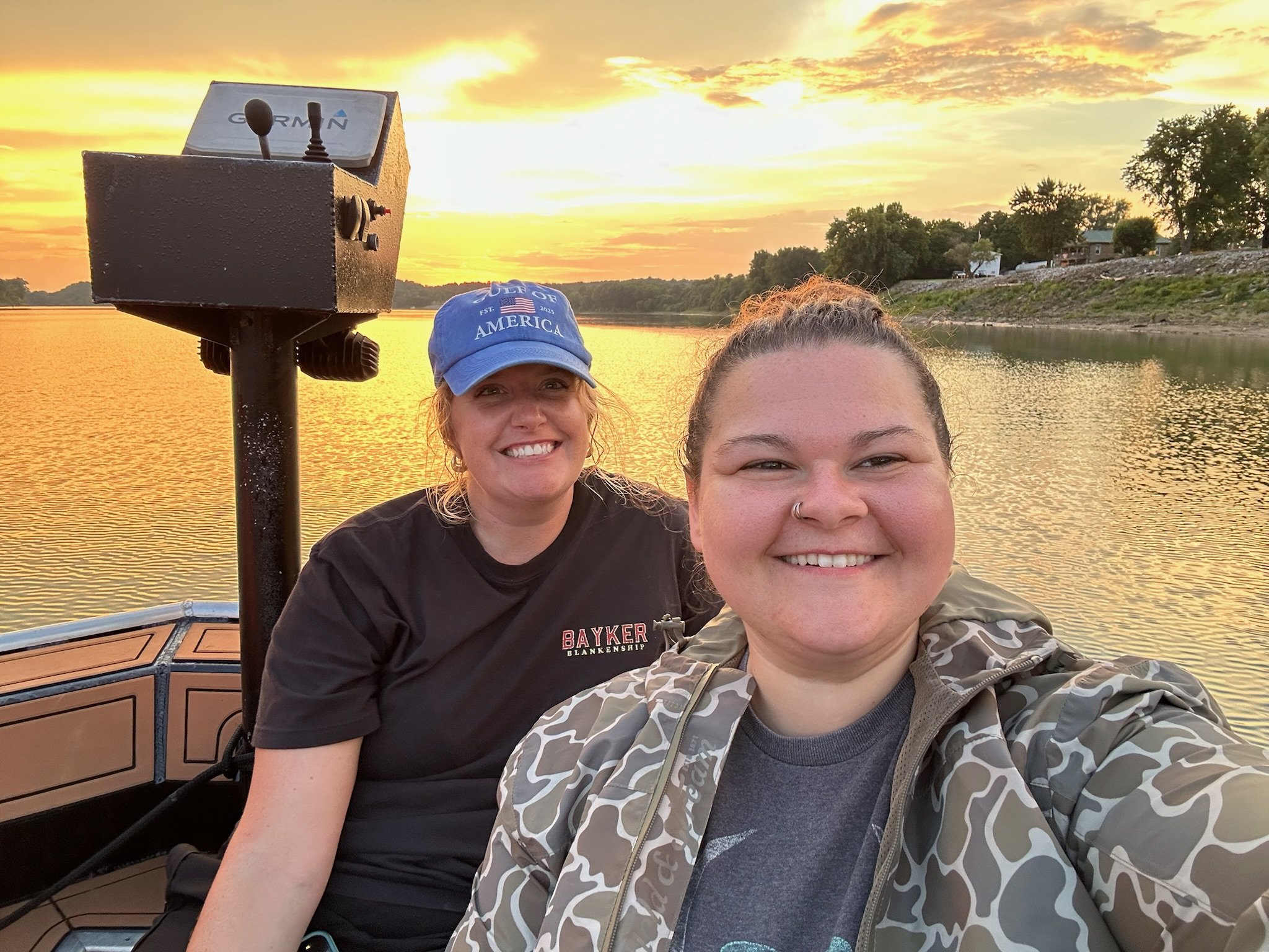 Two women smiling on a boat during sunset, with water and trees in the background.