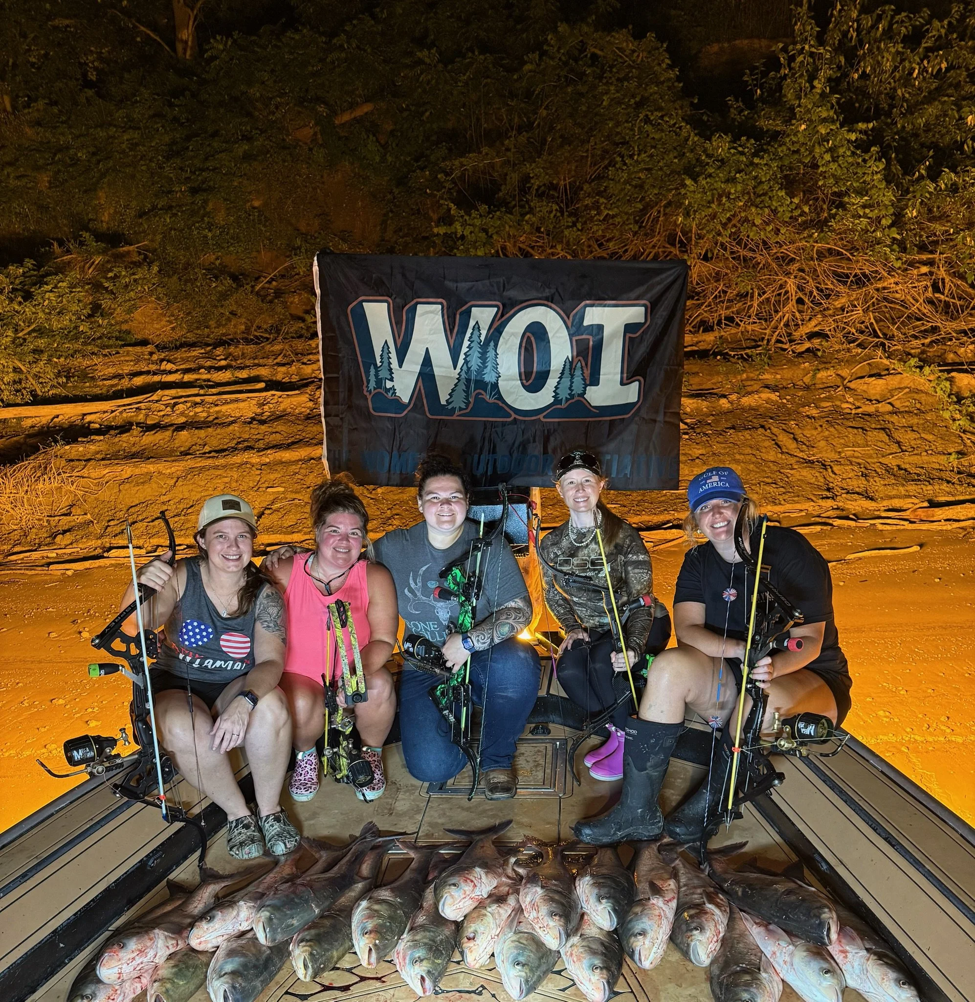 A group of people posing with fishing gear in front of a large catch of fish inside a boat at night. The boat's interior is illuminated, and the background shows a rocky shoreline with dense trees.