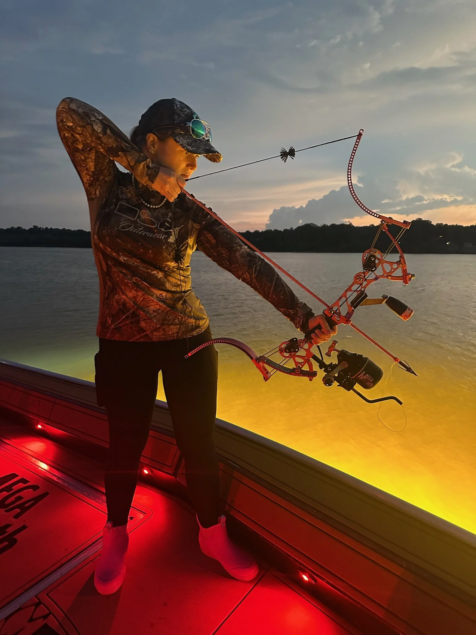 A woman standing on a boat holding a bow and arrow, preparing to shoot, during sunset on a river or lake with a treeline in the background.