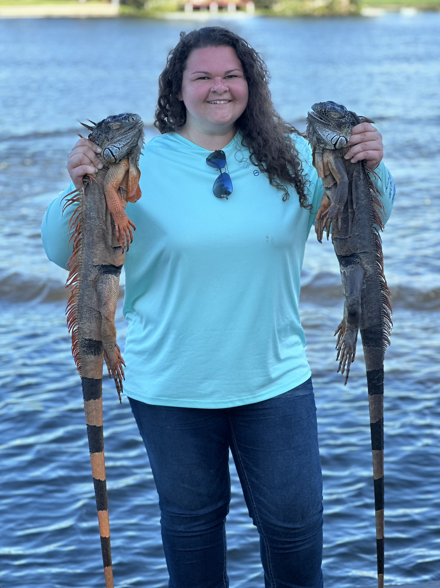 A woman with curly hair, wearing a light blue shirt and jeans, holding two large iguanas near a body of water with greenery in the background.