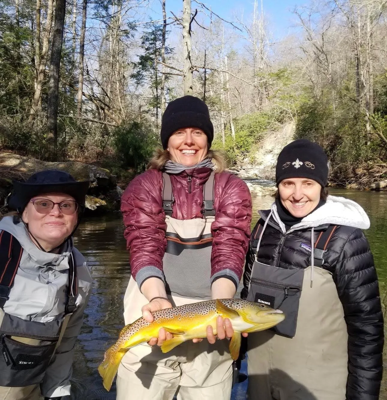 Three women outdoors on a riverbank holding a large, colorful fish, with trees and a waterfall in the background, all dressed in outdoor fishing gear and smiling.