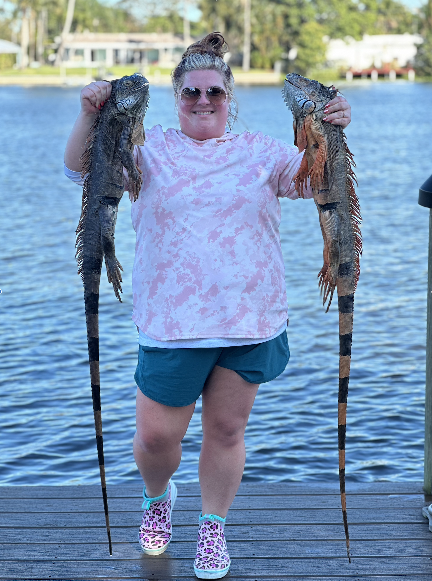 A woman in shorts and pink shirt holding two large iguanas by their tails, standing on a wooden dock by a lake with trees and houses in the background.