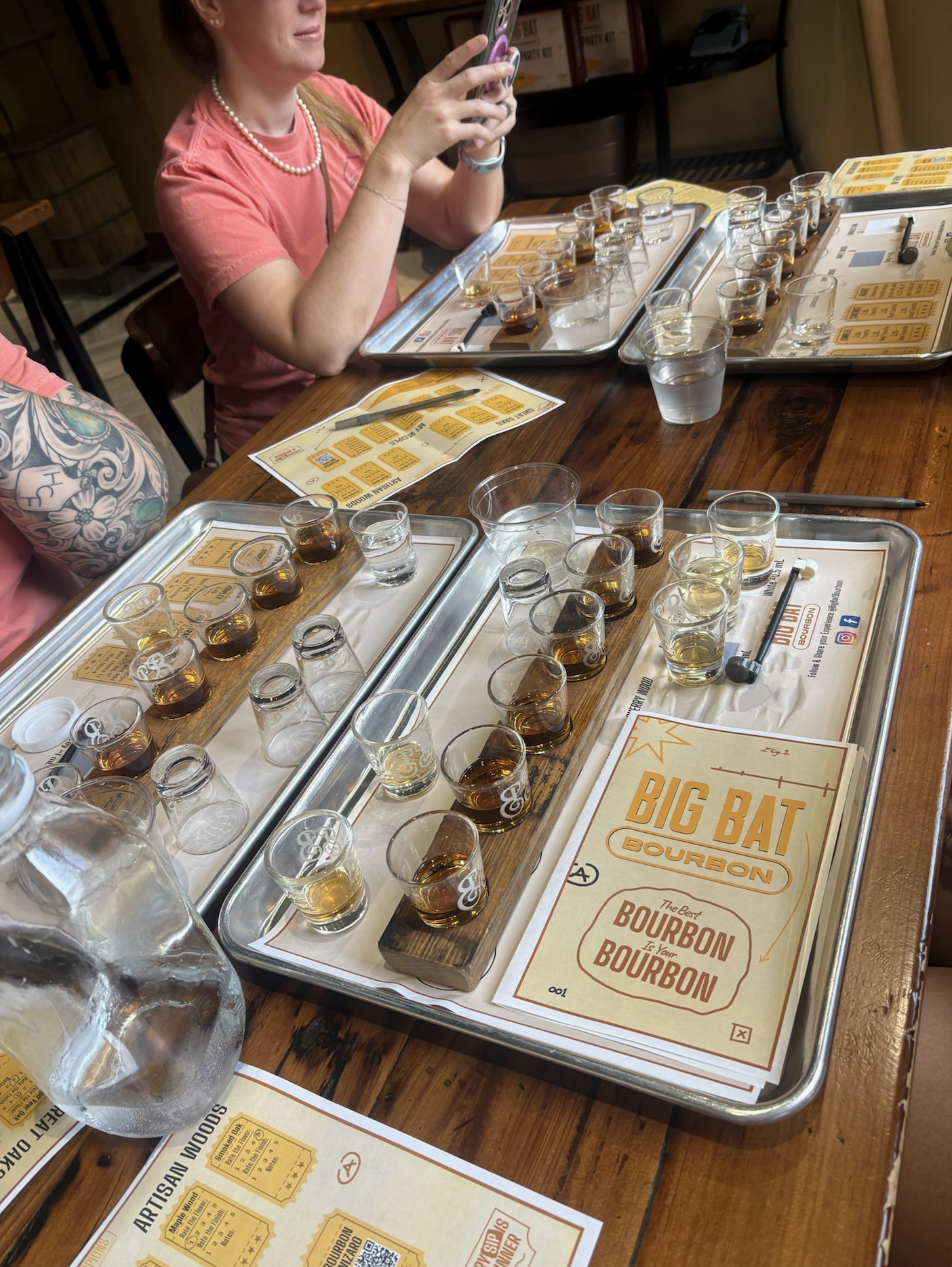 Multiple metal trays with shot glasses filled with bourbon, relighting a cozy tavern environment, with menus and tasting sheets on a wooden table.