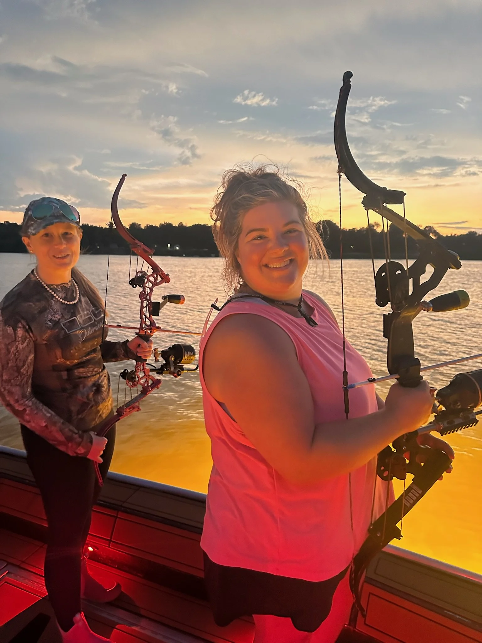Two women smiling at sunset while holding bows and arrows on a boat near a lake.