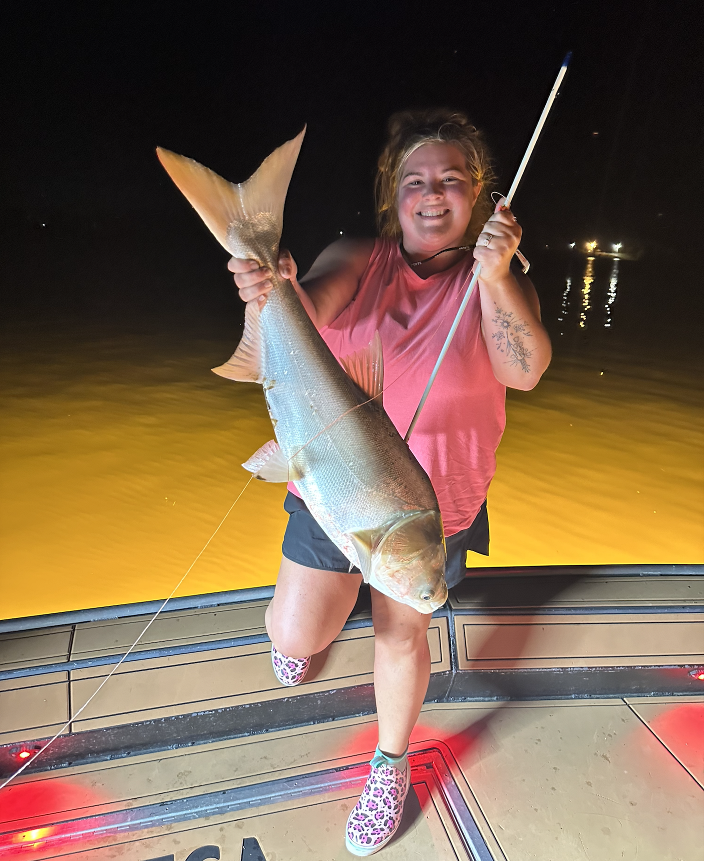 A woman smiling and kneeling on a boat deck during nighttime, holding a large fish with a light-colored body and a wide tail. She is wearing a pink sleeveless shirt, black shorts, and leopard-print shoes. Background shows a body of water with reflections and distant lights.