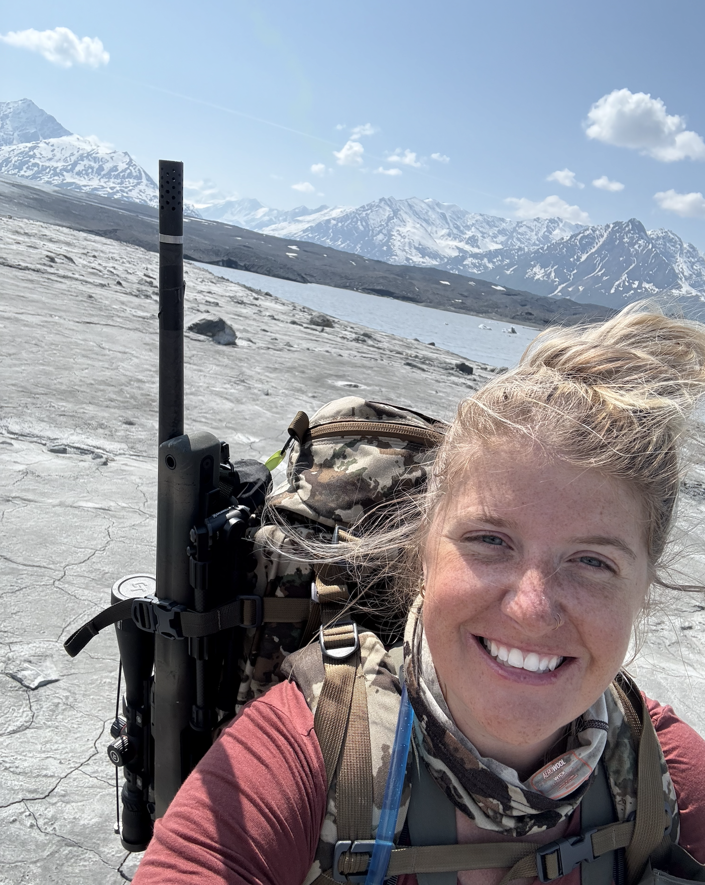 A woman taking a selfie outdoors with snow-capped mountains and a lake in the background. She is smiling, wearing outdoor gear and has long hair.