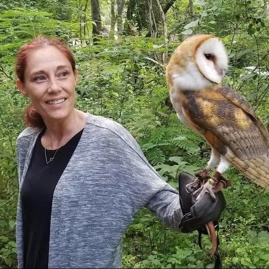 Woman in gray jacket holding a barn owl perched on her gloved hand in a green forest.