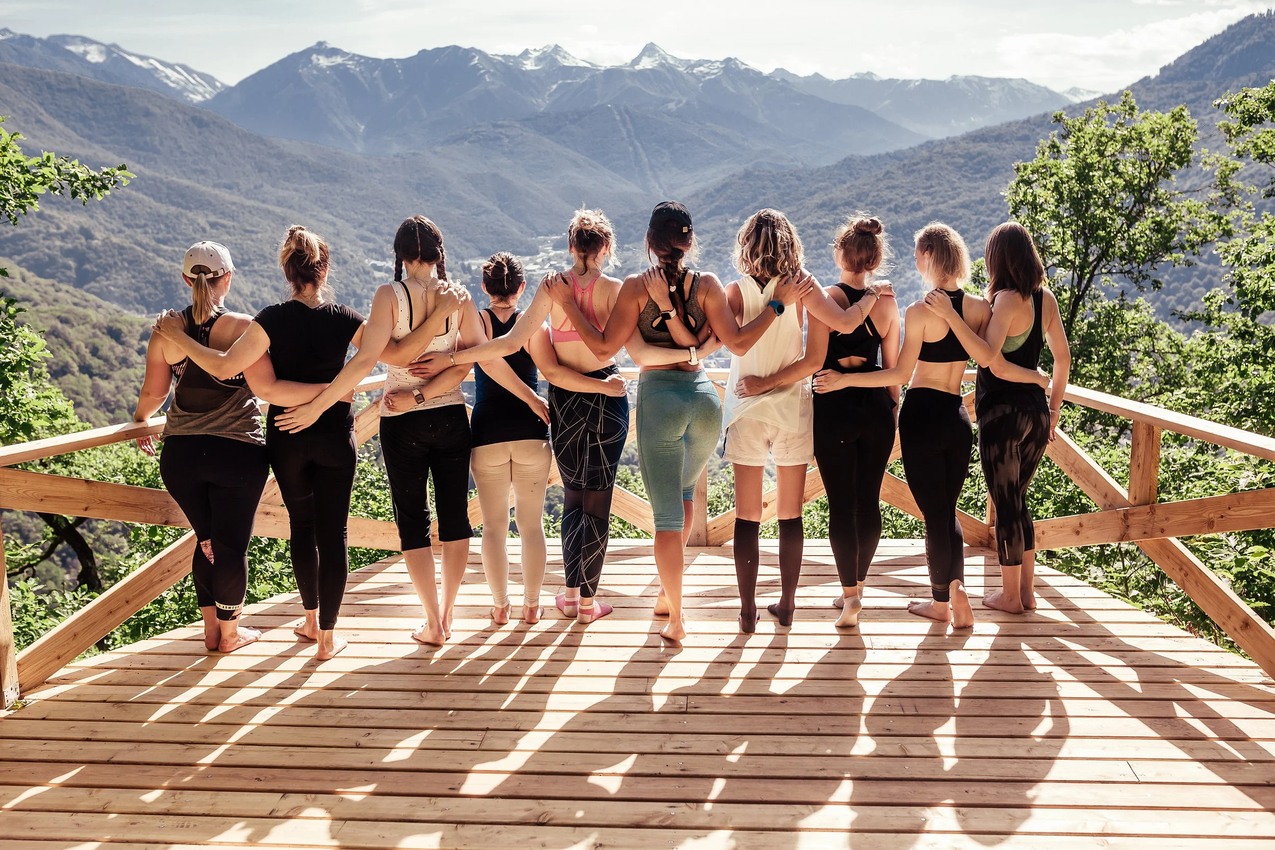 A group of women practicing yoga on a wooden deck outdoors, overlooking a mountain range with snow-capped peaks.