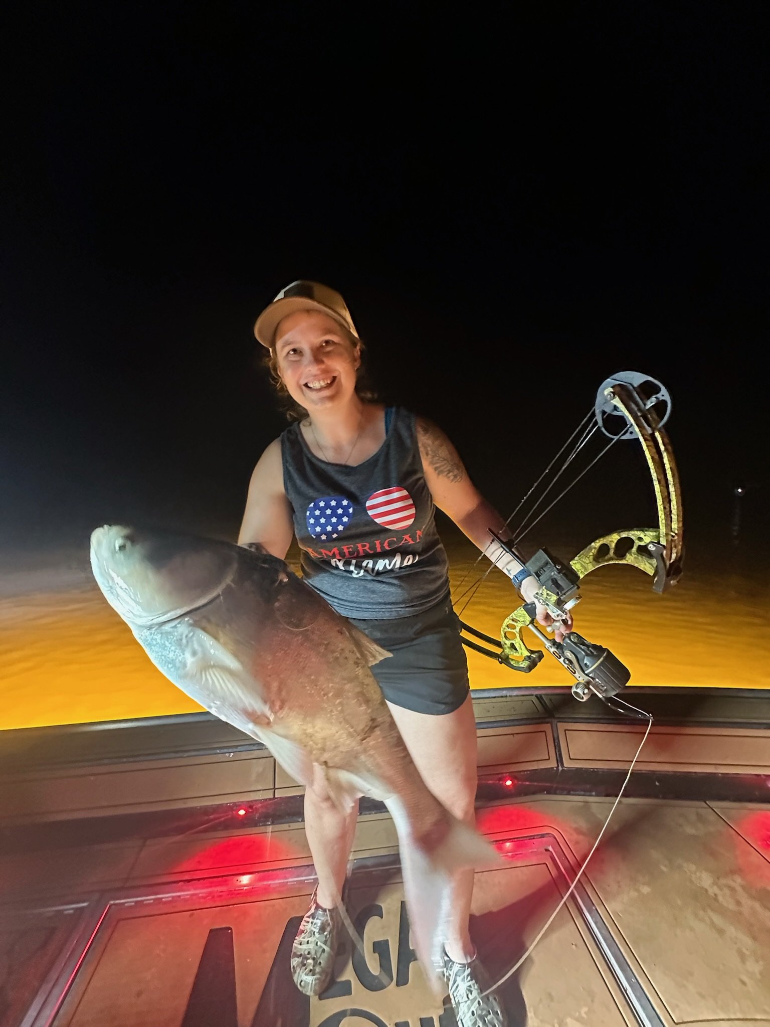 A woman holding a large fish indoors at night, smiling, wearing a cap, tank top, and shorts, with archery equipment visible in the background.