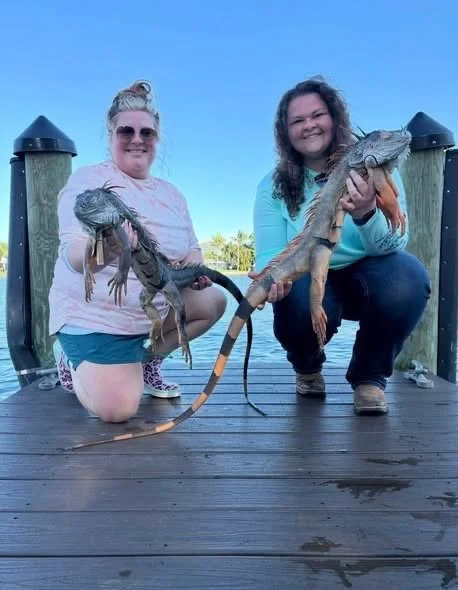 Two women kneeling on a dock holding large alligators, with a blue sky and water in the background.