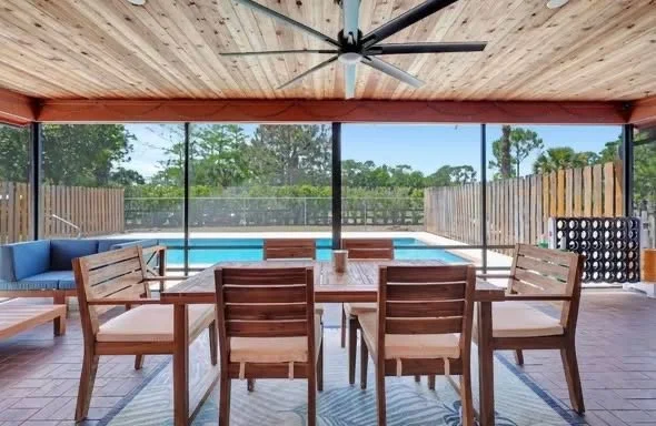 Indoor patio with wooden dining table and chairs, pool, and screened enclosure with sliding glass door, overlooking green trees.