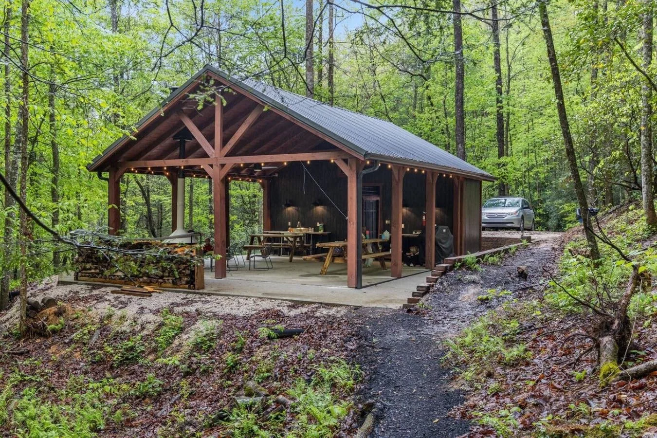 A covered outdoor pavilion in a forest with wooden support beams, string lights, picnic tables, a woodpile, and a parked car on a dirt road.